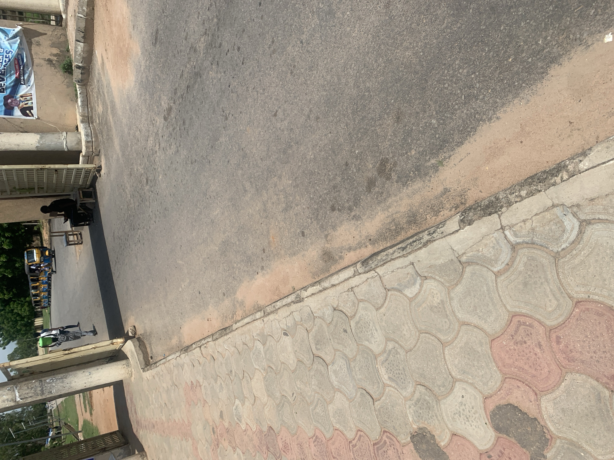 The image depicts an exterior scene on a sunny day, likely in Jalingo, Nigeria. A paved walkway with interlocking concrete tiles, featuring red and gray patterns, runs along the left side of the frame. Adjacent to the walkway is an asphalt road, with a lighter colored sandy area bordering the road and walkway. In the background, a line of yellow and blue auto-rickshaws are parked. A man wearing a gray shirt and trousers carries a green bucket on his head and walks away from the viewer, entering through an open gate. To the right of the gate, a person is seated on a small wooden stool. A banner with an image of a person in graduation attire and text in English is affixed to a building wall. The environment appears to be an urban or semi-urban area, possibly an entrance to an institution or market.