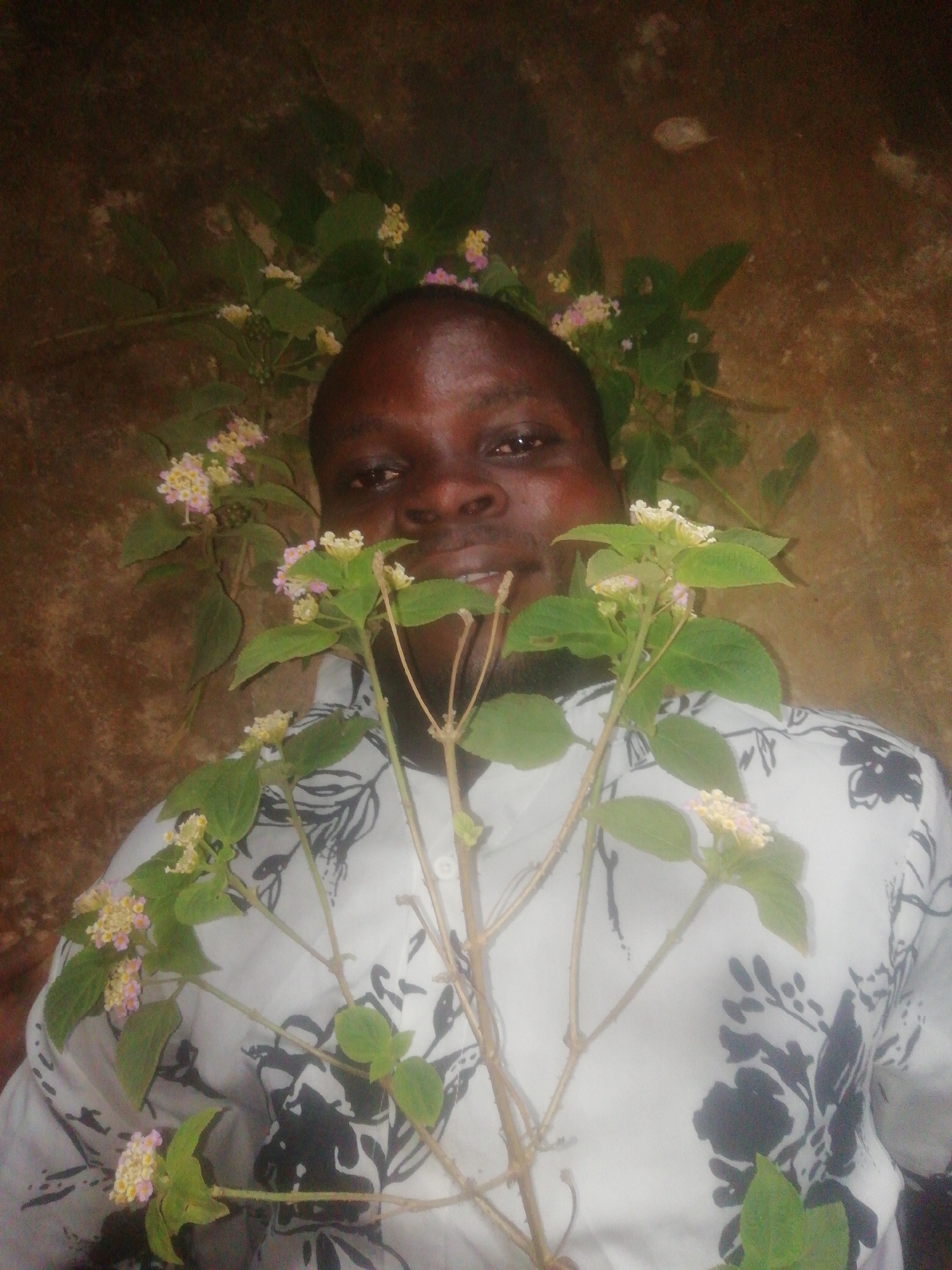 A man lies on a mottled, textured surface, possibly a floor or ground, in an area that is dimly lit. He is of dark complexion and is wearing a white shirt with a black floral pattern. He appears to be holding or be surrounded by branches of a flowering plant with green leaves and clusters of small, multi-colored flowers (pink and yellow). The specific location in the city of Unknown, Unknown is indeterminable from the image provided.