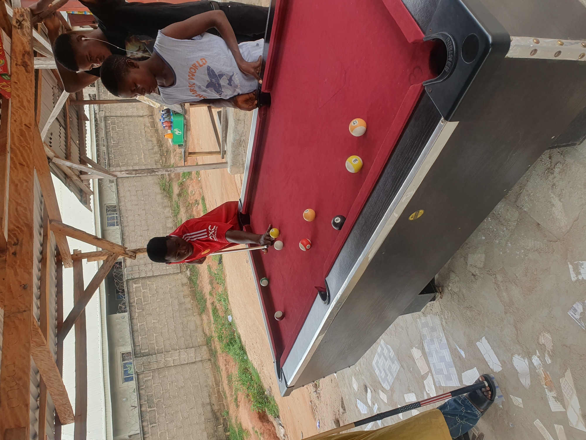 In Lokoja, Nigeria, an outdoor scene features a pool table with red felt and a dark wood exterior. Three young individuals are present. One person, wearing a red and white soccer jersey, is actively playing pool, holding a cue stick and striking a yellow ball. Another person, dressed in a white tank top with "Shark World" printed on it, stands behind the table, observing. A third individual in a black t-shirt stands behind them. The pool table is populated with several billiard balls, including a solid yellow ball, a striped ball with the number 13, a solid yellow ball with the number 9, a solid brown ball with white markings, and a solid black ball with the number 8. The surrounding environment consists of a rough, dirt ground with scattered mosaic tiles. In the background, a brick wall and a simple open-air structure made of wood and corrugated metal roofing provide shelter. A table with various bottles of drinks is visible in the distance. The overall setting suggests a casual recreational area.