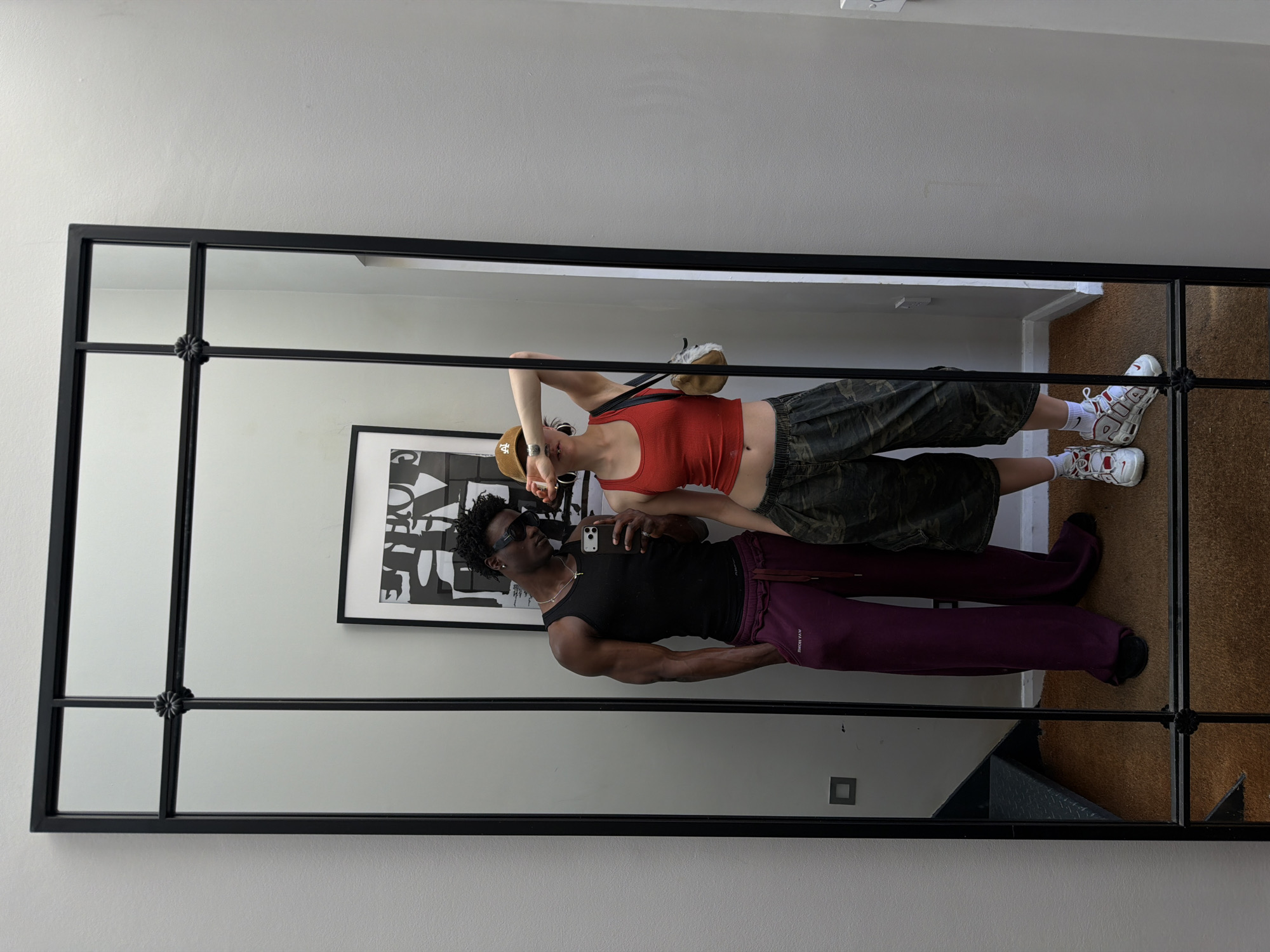 The image depicts a man and woman reflected in a large, rectangular mirror with a black grid frame, mounted on a neutral-colored wall. The woman, positioned higher in the reflection, wears a red tank top, camo pants, white socks, white sneakers, and a tan baseball cap. She has a tan purse slung over her shoulder and is holding a cell phone in her right hand. The man, positioned lower in the reflection, wears a black tank top, purple sweatpants, black socks and black shoes. He wears dark sunglasses. The reflection shows the individuals are standing on a brown carpeted floor in front of the mirror. There is a framed, black and white abstract artwork hanging on the wall behind the individuals. To the right of the mirror’s reflection, part of a wall is visible, alongside a light switch. The location of the image is the city of London, United Kingdom.