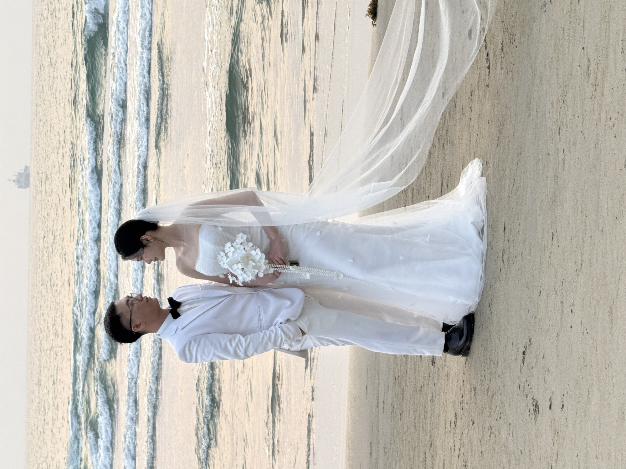 A couple is standing on a beach. The woman is wearing a white wedding dress with a long veil and holding a white bouquet and a pearl necklace. The man is wearing a white suit with a black bowtie. They are standing next to the ocean. The location is likely in Đà Nẵng, Vietnam, given the coastal environment.