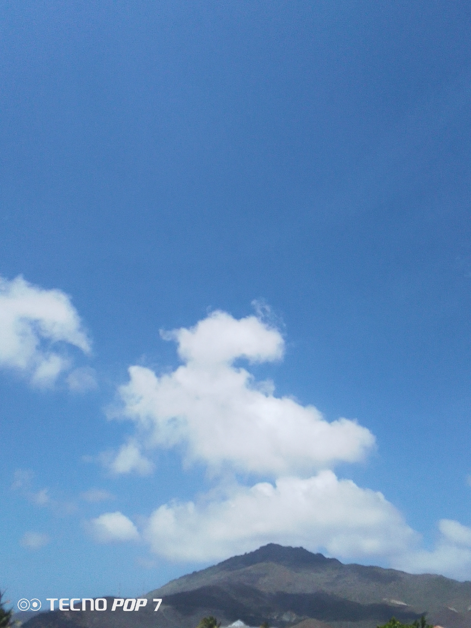 The photograph shows a blue sky with scattered white clouds above a mountain range. The mountains are dark gray-green and have sparse vegetation. The lower portion of the image shows some buildings and greenery, indicating a populated area at the base of the mountains. The text "TECNO POP 7" is visible in the lower left corner. The location is Pedro González, Venezuela.