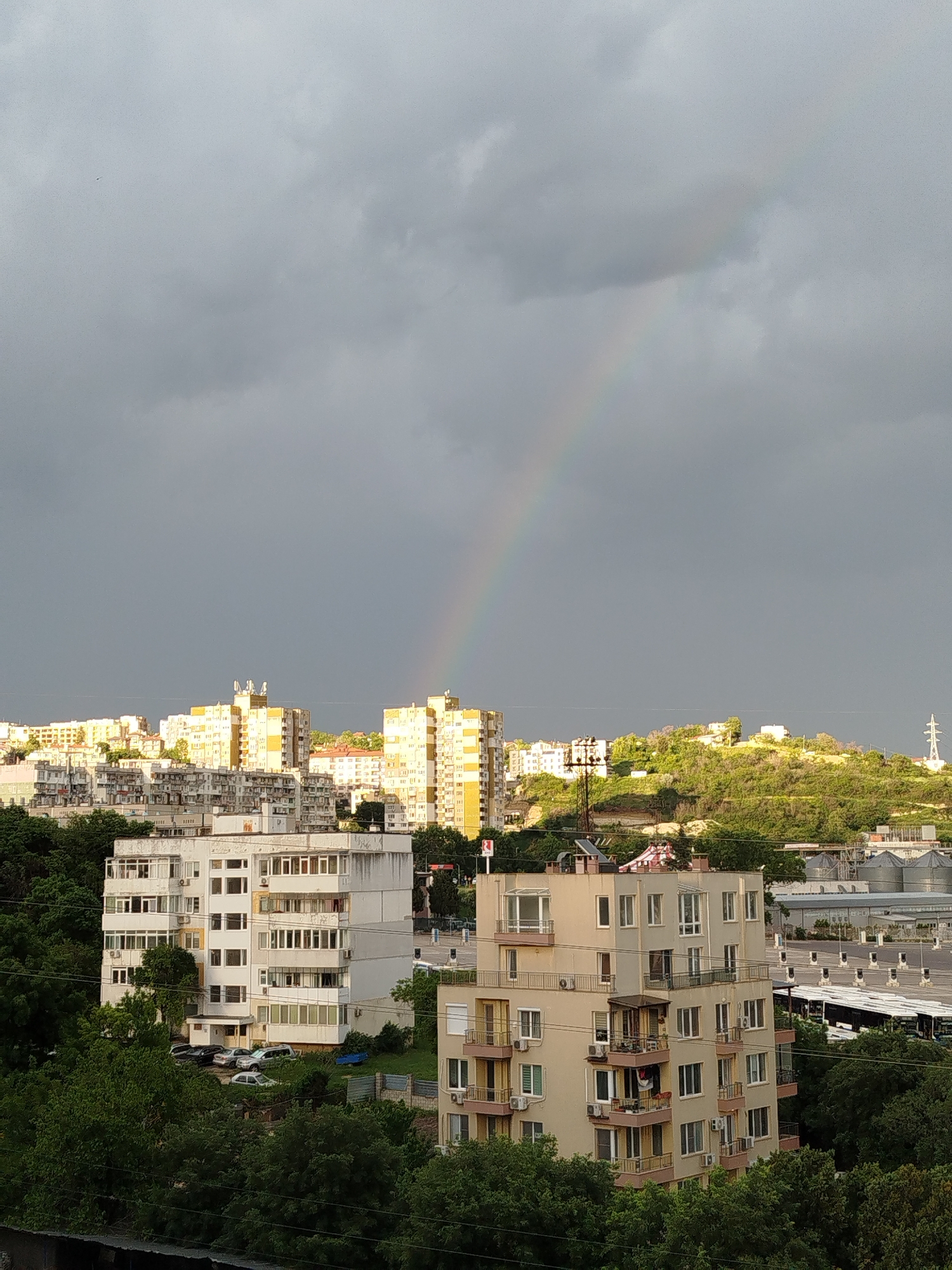 The image captures a view of Varna, Bulgaria under a stormy sky with a faint rainbow arc. The foreground features residential buildings, primarily of white and beige coloring, nestled amidst green trees. In the background, taller yellow and white apartment blocks stand out against the horizon. Beyond the buildings, a green hill rises. To the right, an industrial area or parking lot is visible with several silos or storage tanks and buses. The sky is predominantly gray with darker storm clouds dominating the upper portion of the frame.