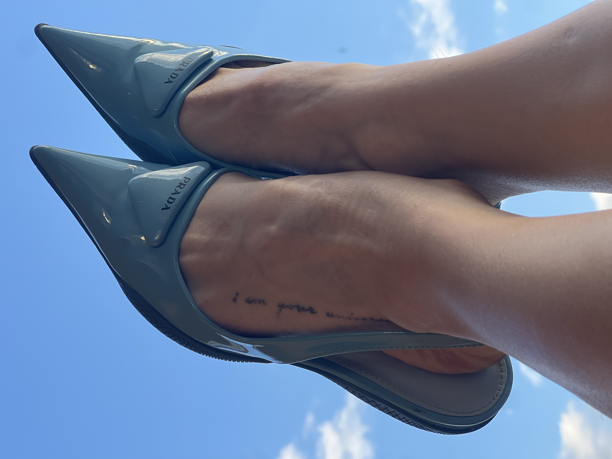 The image shows a low-angle, close-up shot of two feet wearing light blue Prada mules against a bright blue sky with scattered white clouds. The feet are positioned with the soles facing upward, revealing the interior of the shoes which are also light blue. The shoes have pointed toes and a subtle logo, "PRADA," printed on the exterior near the heel. On the left foot, a small, vertical tattoo is visible on the ankle, appearing to be in script. The skin tone is light to medium. The image does not contain any recognizable landmarks or elements that would suggest a specific location in Saint Petersburg, Russia.