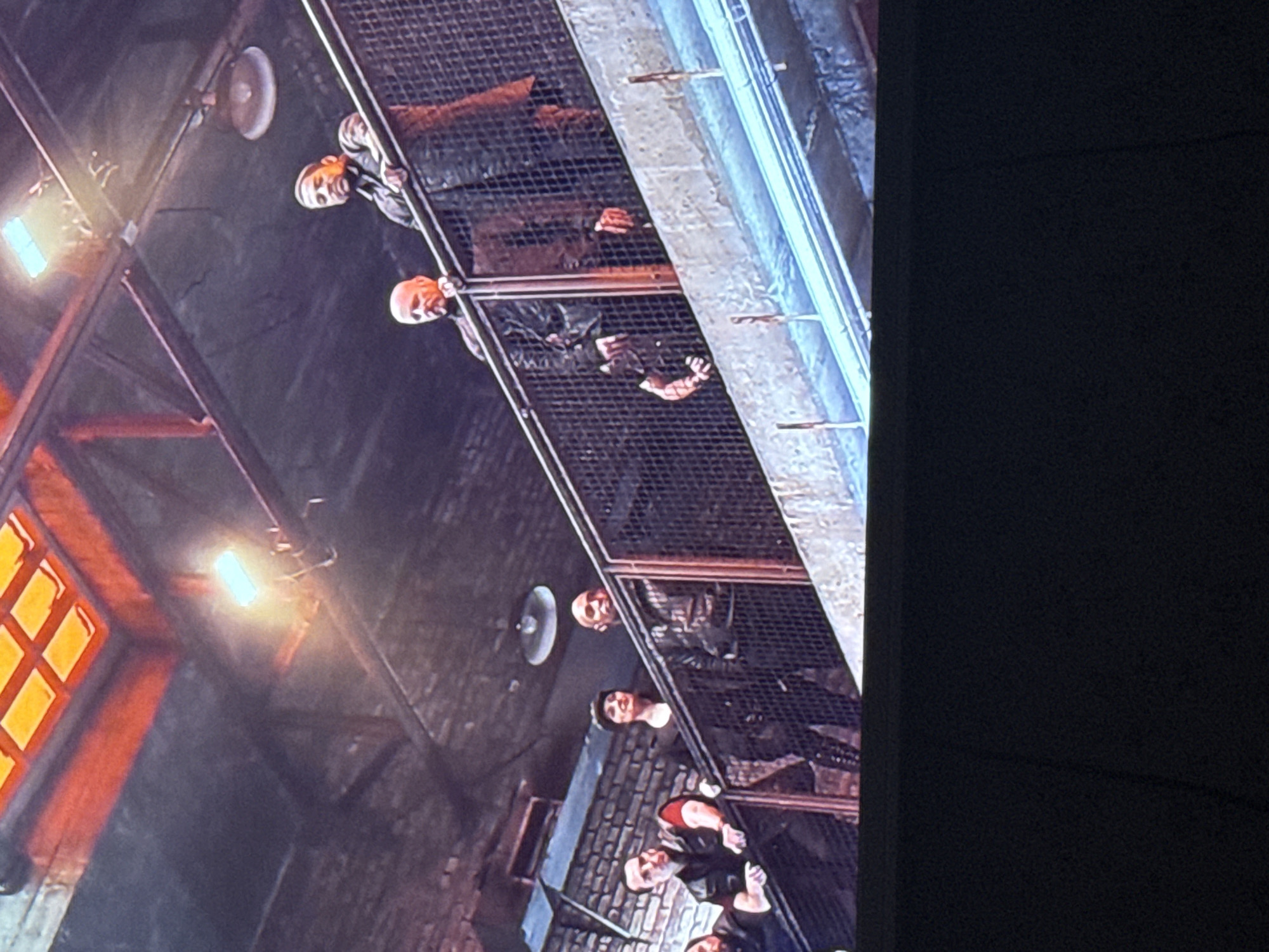 The image depicts a group of people standing on a metal grate above what appears to be an alleyway or industrial area. The scene is dimly lit by overhead lights and the warm glow from a distant window with a grid pattern. The individuals are looking down, their expressions and actions not readily discernible. The environment suggests an urban or industrial location, with brick walls and overhead fixtures contributing to the atmosphere. The city of Mytishchi, Russia is not verifiable from the image provided.