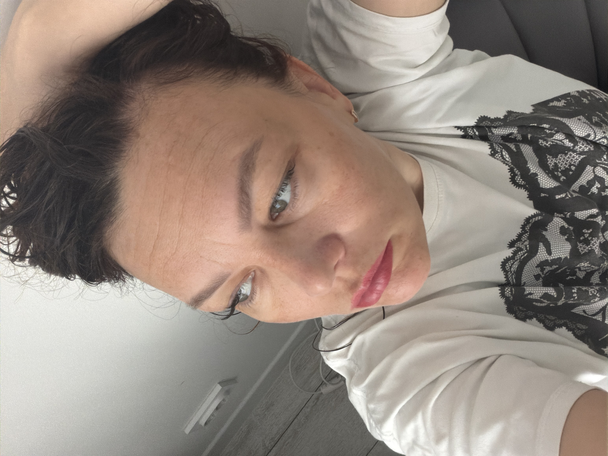 A selfie of a woman with dark, curly hair, light skin, and green eyes. She is wearing a white T-shirt with a black lace-like design on the front. She appears to be indoors, based on the presence of a white wall and a glimpse of flooring. The image is taken from a slightly above and angled perspective. The location is unknown.