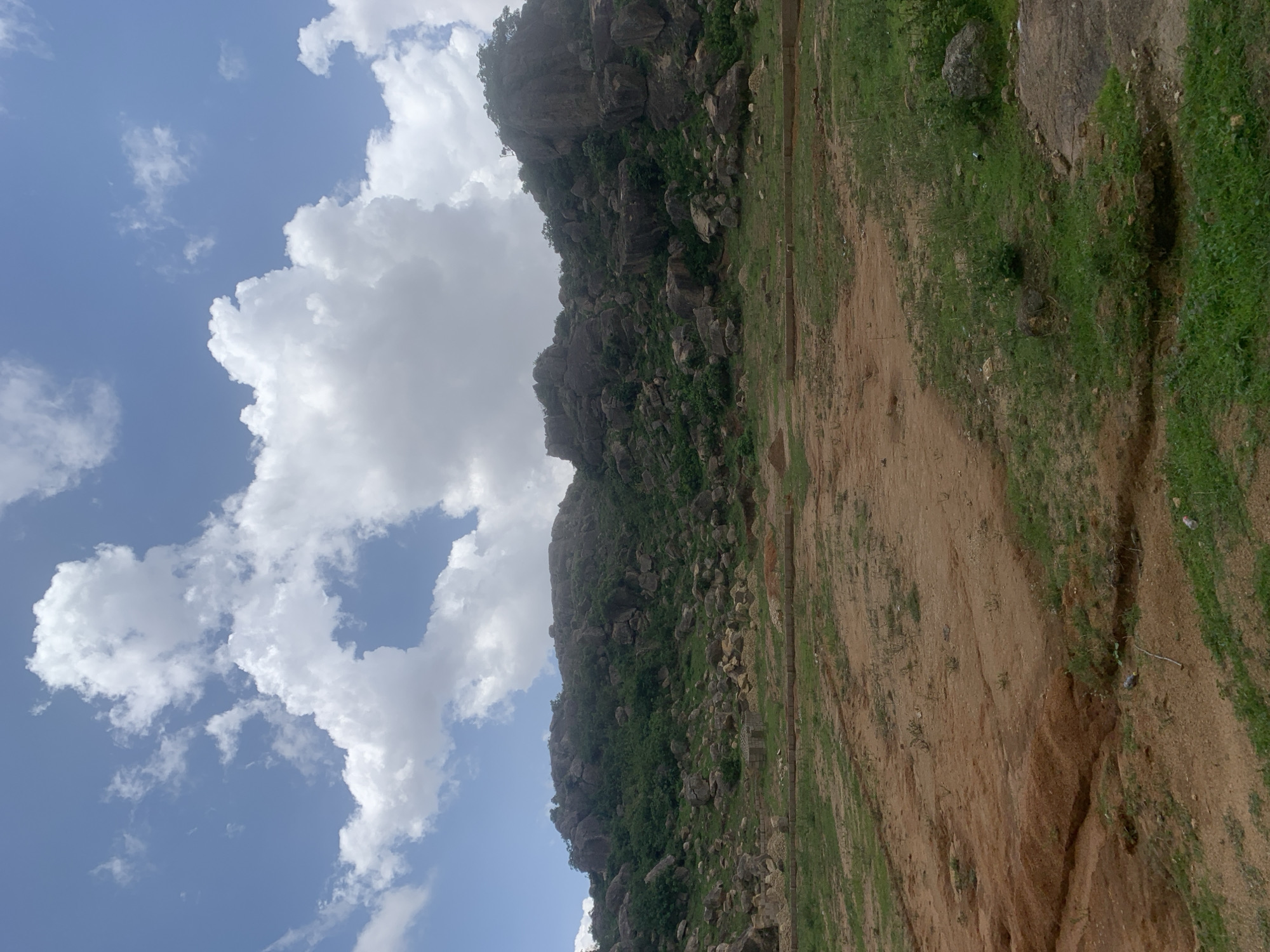 The image depicts a natural landscape with a rocky hillside in the background. The hillside is covered in vegetation and large boulders. In the foreground, there is an area of uneven, dry earth interspersed with patches of green grass. There are also some low walls or construction elements visible in the mid-ground. The sky above is blue with scattered white clouds. The scene appears to be an undeveloped or partially developed area, possibly on the outskirts of a settlement. The location is identified as Barakin Akawo, Nigeria. There are no people or visible signs of recent activity in the image.