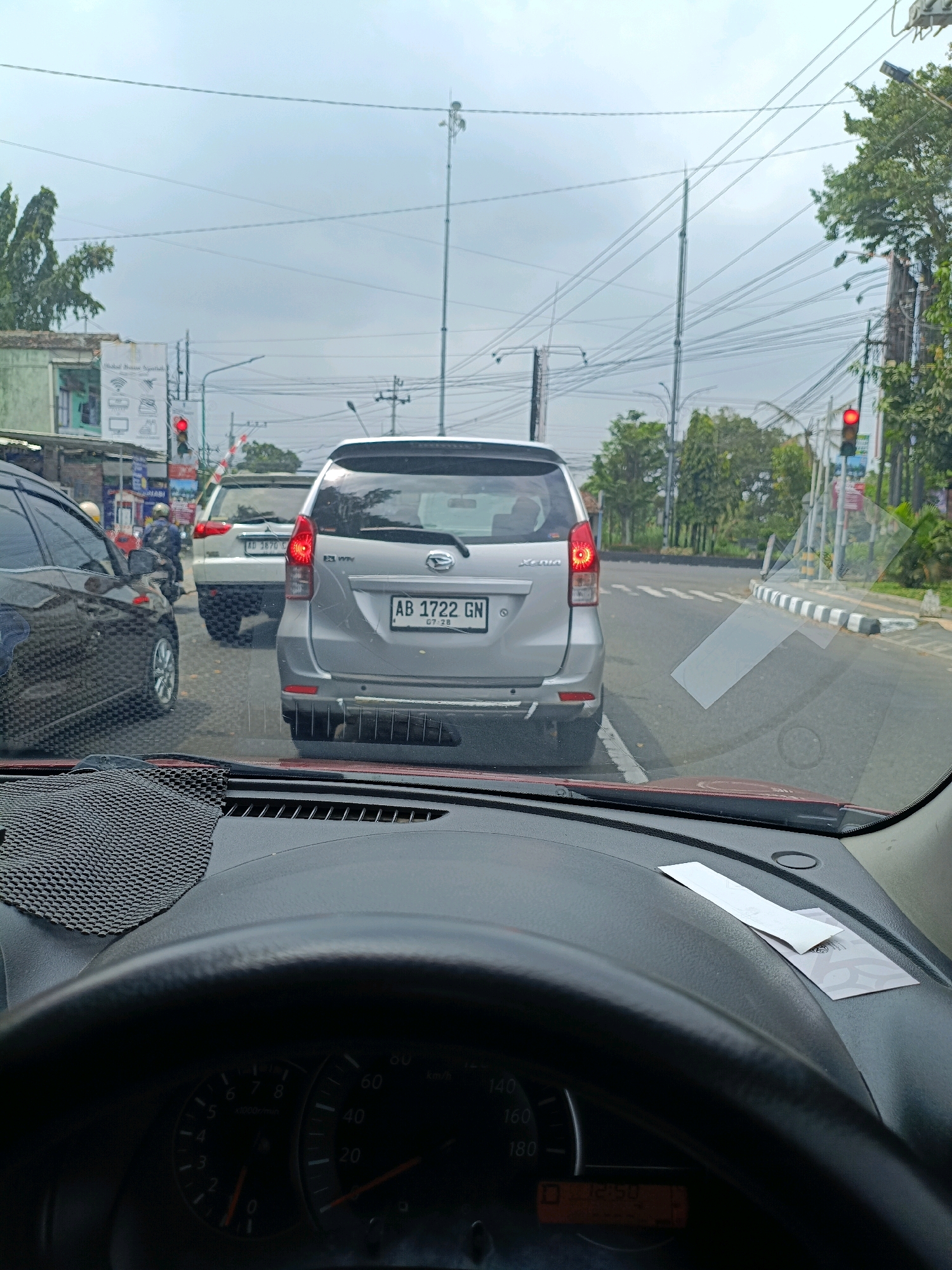 The image shows a street scene in Sleman, Indonesia, from the perspective of a driver inside a car. The car's dashboard, steering wheel, and windshield are visible in the foreground, partially obscuring the view of the road ahead. Several vehicles are stopped on the road, indicating traffic congestion. A silver minivan with the license plate "AB 1722 GN" is prominent in the center of the frame, with its brake lights illuminated. To its left, a white SUV with license plate "AB 3870 G" is also stopped. A dark-colored car is visible to the left of the minivan. A traffic light, showing red, is visible in the background, along with power lines and utility poles. Trees and buildings line the sides of the street. The sky is overcast.