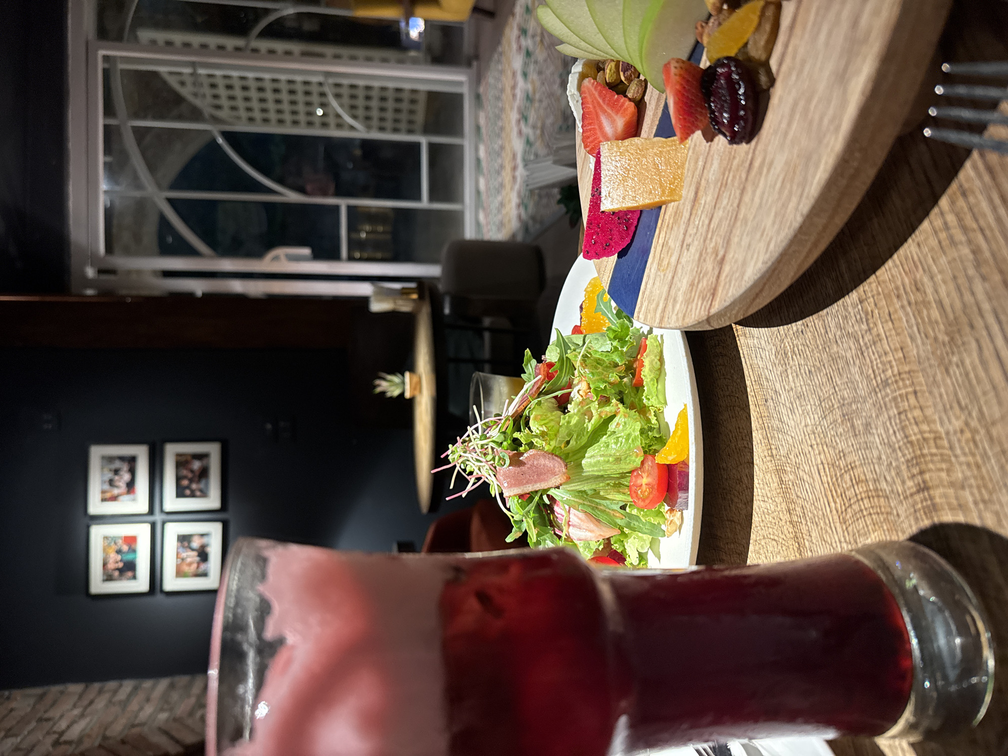 This media file shows a close-up view of a meal, likely at a restaurant or home in Hanoi, Vietnam. In the foreground, a tall glass filled with a frothy, pink and dark red beverage is prominently featured. The drink appears to be layered, with a lighter pink foam on top and a darker liquid below.
Behind the drink, a white plate holds a vibrant salad. The salad consists of fresh green lettuce, sliced tomatoes, orange segments, and pieces of what appears to be cured meat. Delicate microgreens are also visible, adding a touch of freshness. To the right of the salad is a wooden board topped with a fruit and cheese platter. This platter includes sliced green apples, strawberries, a piece of orange-colored cheese, dragon fruit slices, dried fruit, and nuts. A fork rests on the edge of the wooden board.
In the background, the setting appears to be an indoor space with ambient lighting, suggesting it might be evening. A window with white latticework is visible, offering a glimpse of an outdoor area or another part of the establishment. Further in the background, on a dark wall, four framed pictures are hung in a square arrangement. The overall scene conveys a relaxed dining experience.