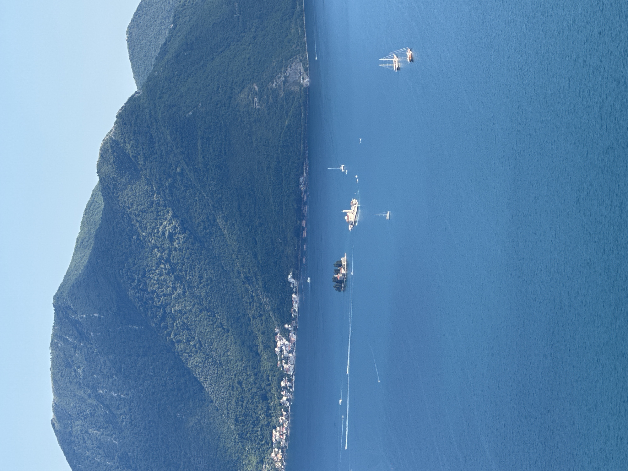 This is an aerial, wide-angle, eye-level shot of a bay with a town and islands. The foreground is dominated by the deep blue water of the bay, with light rippling on its surface. Several boats are visible, including sailboats with white sails and smaller speedboats leaving white wakes. In the middle ground, two small islands are present. The larger island on the left has several buildings with red roofs clustered together. The smaller island to its right has a single building with a red roof. Along the shoreline in the background, a town with many buildings featuring red roofs lines the edge of the water. Behind the town and the bay, a lush, green, mountainous landscape rises steeply, its slopes covered in dense vegetation. The sky above is a clear, pale blue. This scene appears to be located in the area of Strp, Montenegro, known for its scenic bay and historic islands.