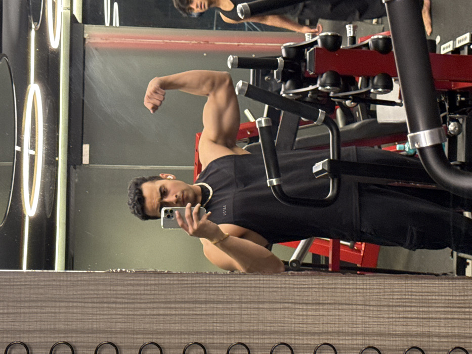 A male individual, identified as being in a gym in Kolkata, India, is captured in a mirror selfie. He is positioned centrally, wearing a black sleeveless tank top with a "MAN" logo, black cargo pants, a gold-colored chain necklace, and a gold-colored bracelet on his left wrist. A white wireless earbud is visible in his right ear. He holds a silver-colored smartphone in his left hand, angled to photograph his reflection, and is flexing his right bicep with a neutral facial expression. Behind him, reflected in the mirror, another male individual with dark curly hair is partially visible to the right, also wearing a black sleeveless top. The gym environment includes black and red weight training equipment; a machine with black handles and silver accents is in the foreground, and a red framed piece of equipment is in the midground. Overhead circular fluorescent lighting fixtures are reflected on the mirror's surface. A blue water bottle is visible on the floor near the red equipment. To the left of the mirror, a textured vertical panel features several dark circular rings.