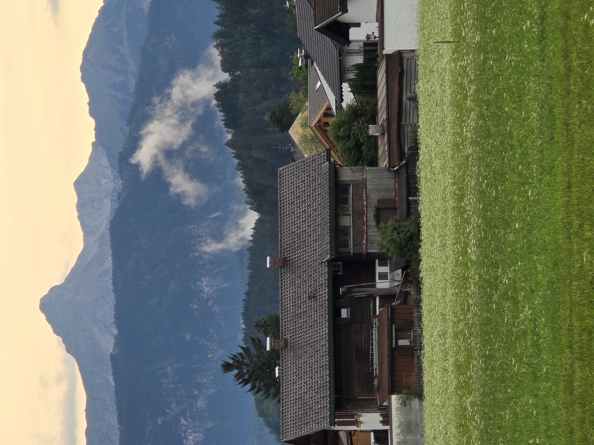 The image depicts a scenic mountain village in Garmisch-Partenkirchen, Germany. The composition is dominated by a towering, rugged mountain range on the left, bathed in soft, yellowish light, suggesting either dawn or dusk. Wispy clouds are scattered across the mountain slopes, adding depth and texture.
In the mid-ground, several traditional wooden houses with tiled roofs are nestled on the mountainside. These buildings exhibit architectural features typical of the Bavarian Alps, with dark wood exteriors and pitched roofs. Some houses have visible windows and chimneys. A vibrant green meadow, dotted with small white wildflowers, stretches across the right side of the frame, creating a stark contrast with the darker hues of the mountains and buildings. The meadow appears lush and well-maintained.
The scene captures a tranquil moment in a rural alpine setting. The overall impression is one of natural beauty and peaceful rural life. No people are visible in the image, and no text is present. The time of day is likely either early morning or late evening due to the warm, diffused lighting and the soft shadows. The weather appears calm, with only light clouds present.