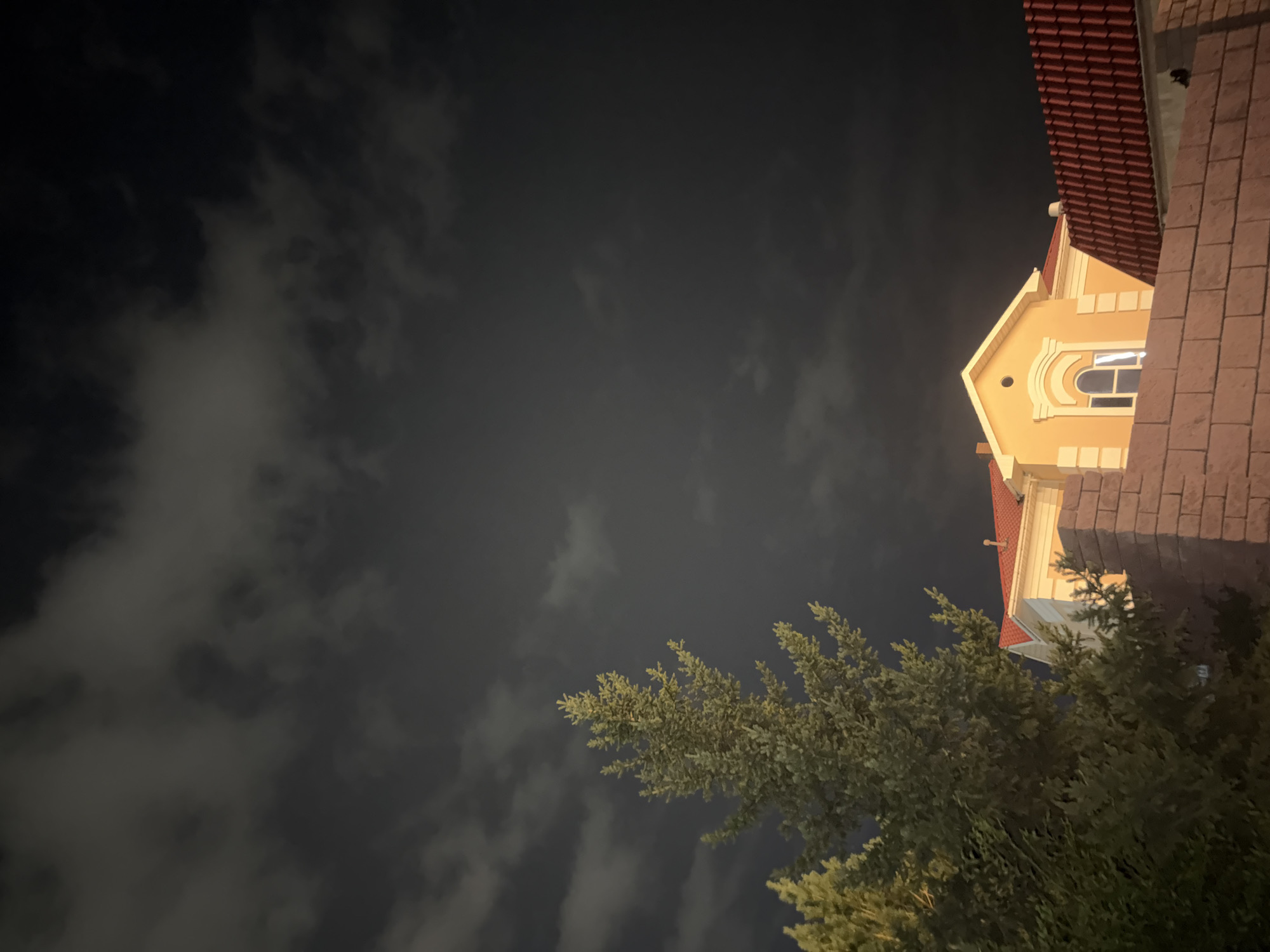 This image depicts a nighttime scene in Pavlodar, Kazakhstan, with a focus on a residential building. In the foreground, a brick wall separates the viewer from the property. Behind the wall, the roof of a house with red tiles is visible. Further back, a light-yellow house with white trim is illuminated, featuring a prominent arched window. To the left of the house, a tall coniferous tree stands, its branches illuminated by an unseen light source. The sky above is dark and filled with cloudy formations, suggesting an overcast or stormy night. No people are visible in the frame.