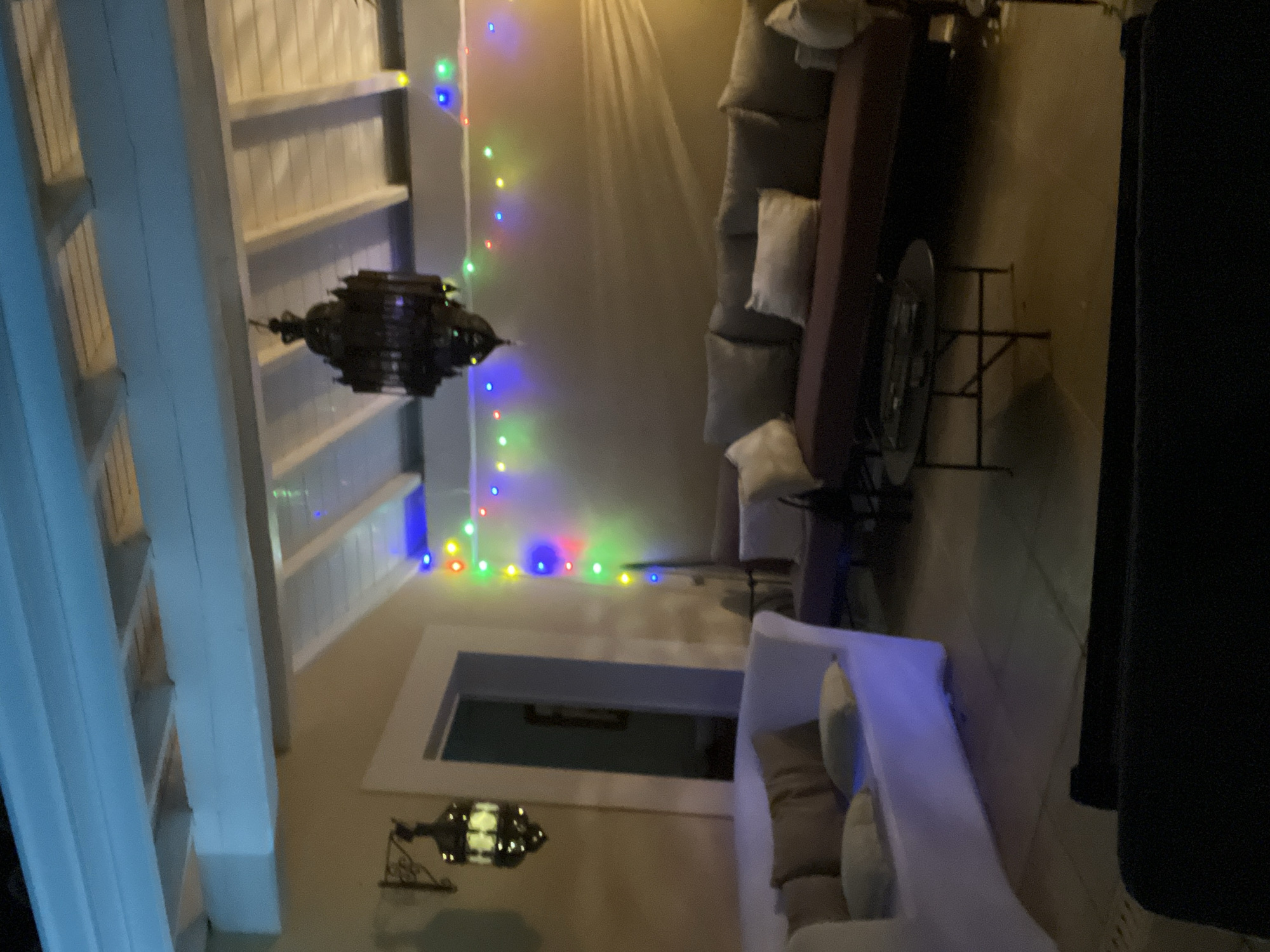 This is a nighttime, medium-shot, eye-level, outdoor photograph of a covered patio or terrace area. The scene is illuminated by a string of colorful lights draped along the ceiling beams and two ornate hanging lanterns. In the foreground, there is a modern-looking white sofa with beige cushions. Behind it, a dark-colored coffee table with a metal base sits on a tiled floor. Further back, a long, dark-colored sofa with multiple cushions is visible. The walls are a neutral beige color, and the ceiling features exposed white beams with slatted wood panels above. A doorway leading to an interior space is partially visible on the left. The overall atmosphere is relaxed and inviting, suggesting a comfortable outdoor living space. The location is identified as Saint-Galmier, France.