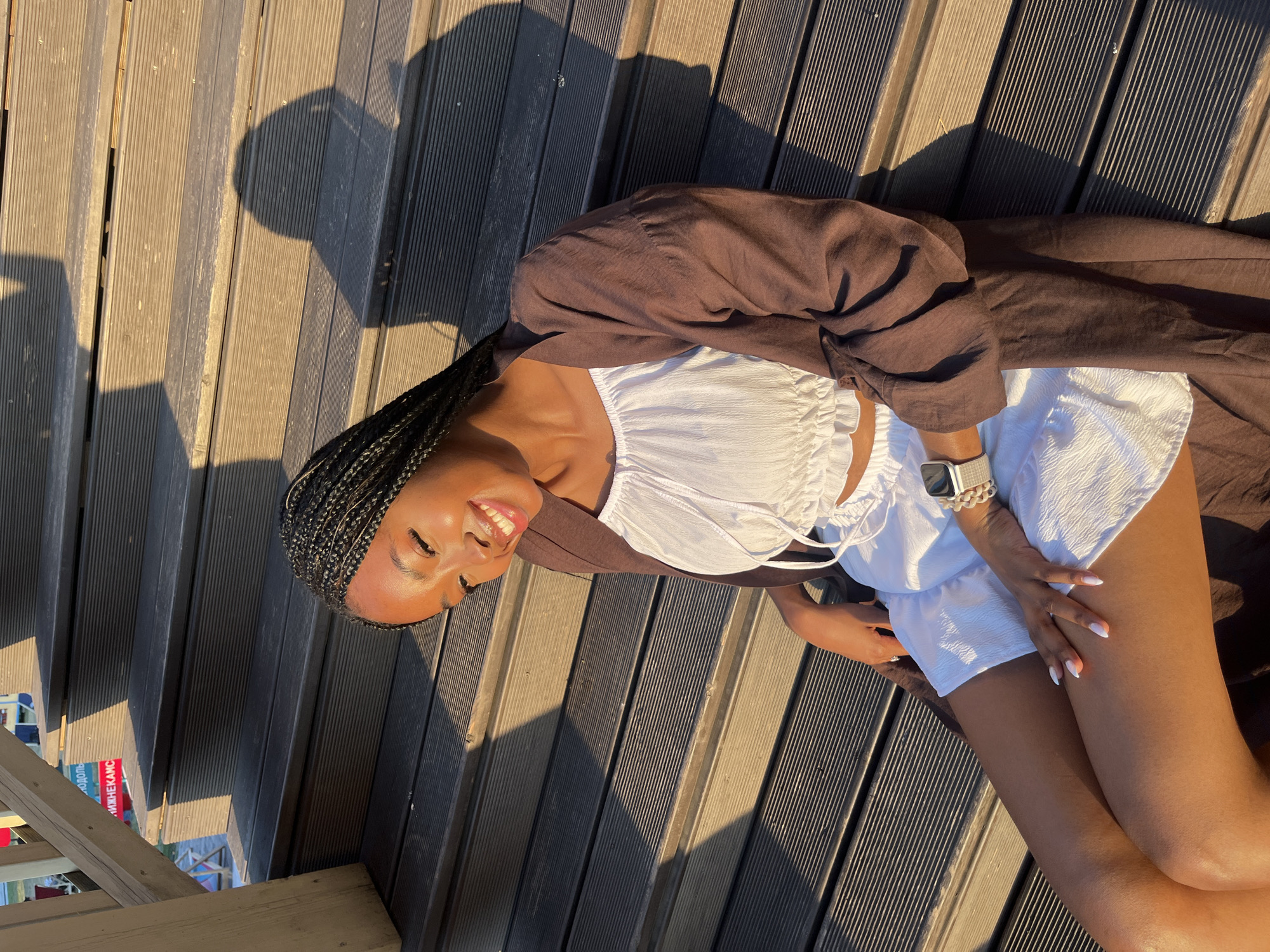 A young Black woman with braided hair and a white crop top and white shorts sits on wooden stairs in Kazan, Russia. She is wearing a brown cardigan and a silver watch. The sun is setting, casting a shadow on the stairs. A sign with Russian text is visible in the background.