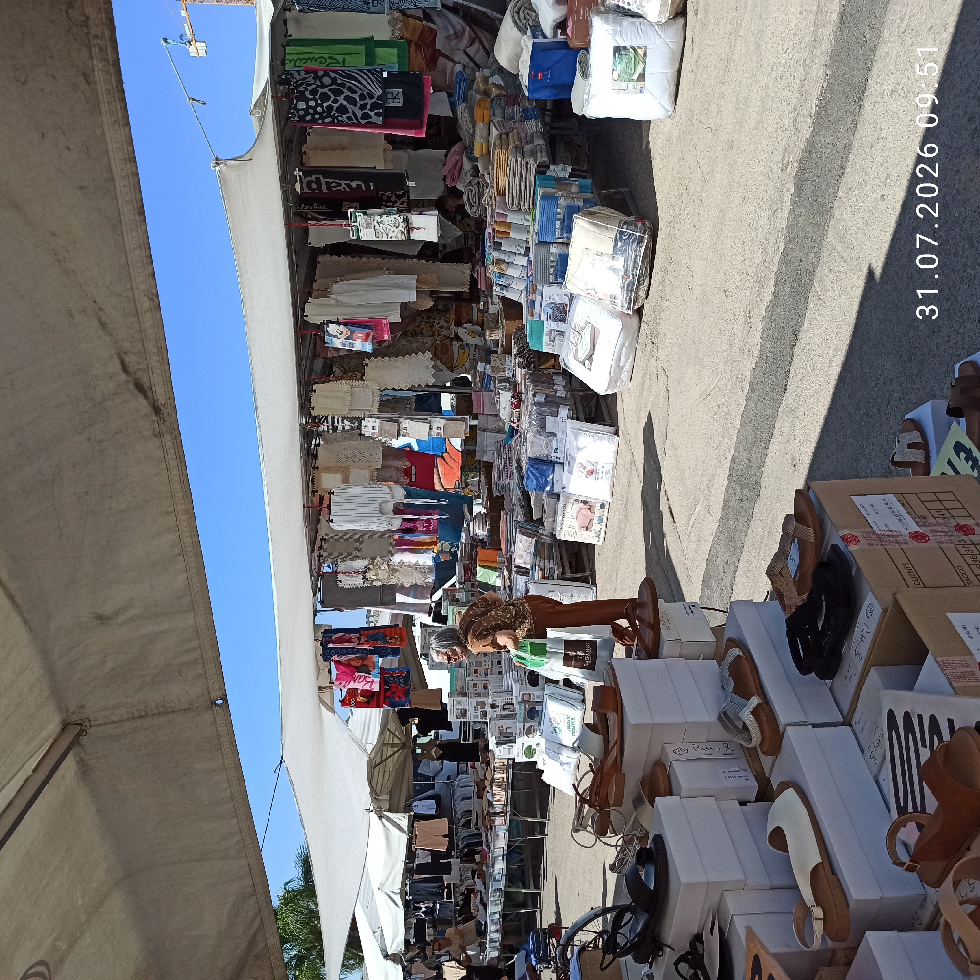 This is a wide, eye-level, outdoor shot of a street market in Modugno, Italy. The market is covered by white awnings and stalls draped with various textiles, including towels, bedding, and clothing. In the foreground, several pairs of women's sandals are displayed on white boxes and cardboard. A woman in a brown top and pants, carrying a shopping bag, is seen walking through the market. Behind her, a man is visible behind a stall. The ground is paved with asphalt and concrete, with some shadows cast by the awnings and objects. The sky is clear and blue. A construction crane is visible in the background. The timestamp "31.07.2026 09:51" is superimposed on the bottom right of the image.