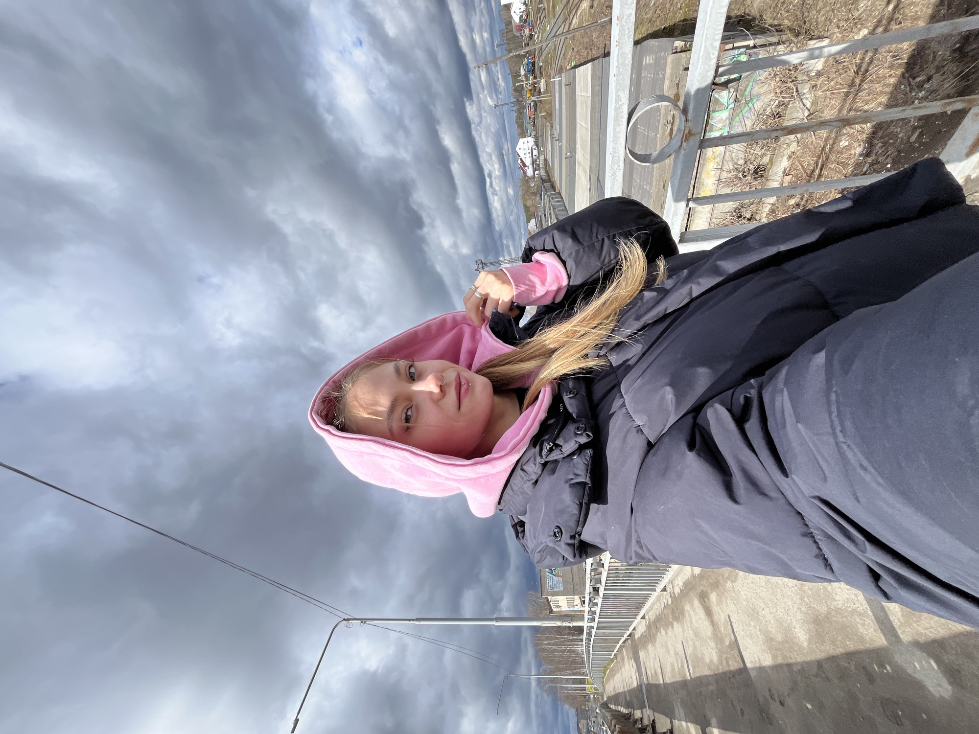 A young woman in a pink hooded sweatshirt and dark blue puffer jacket takes a selfie on a bridge in Chaikovsky, Russia. She is standing next to the bridge's railing, with a cloudy sky in the background. A railway and some industrial buildings can be seen in the distance.