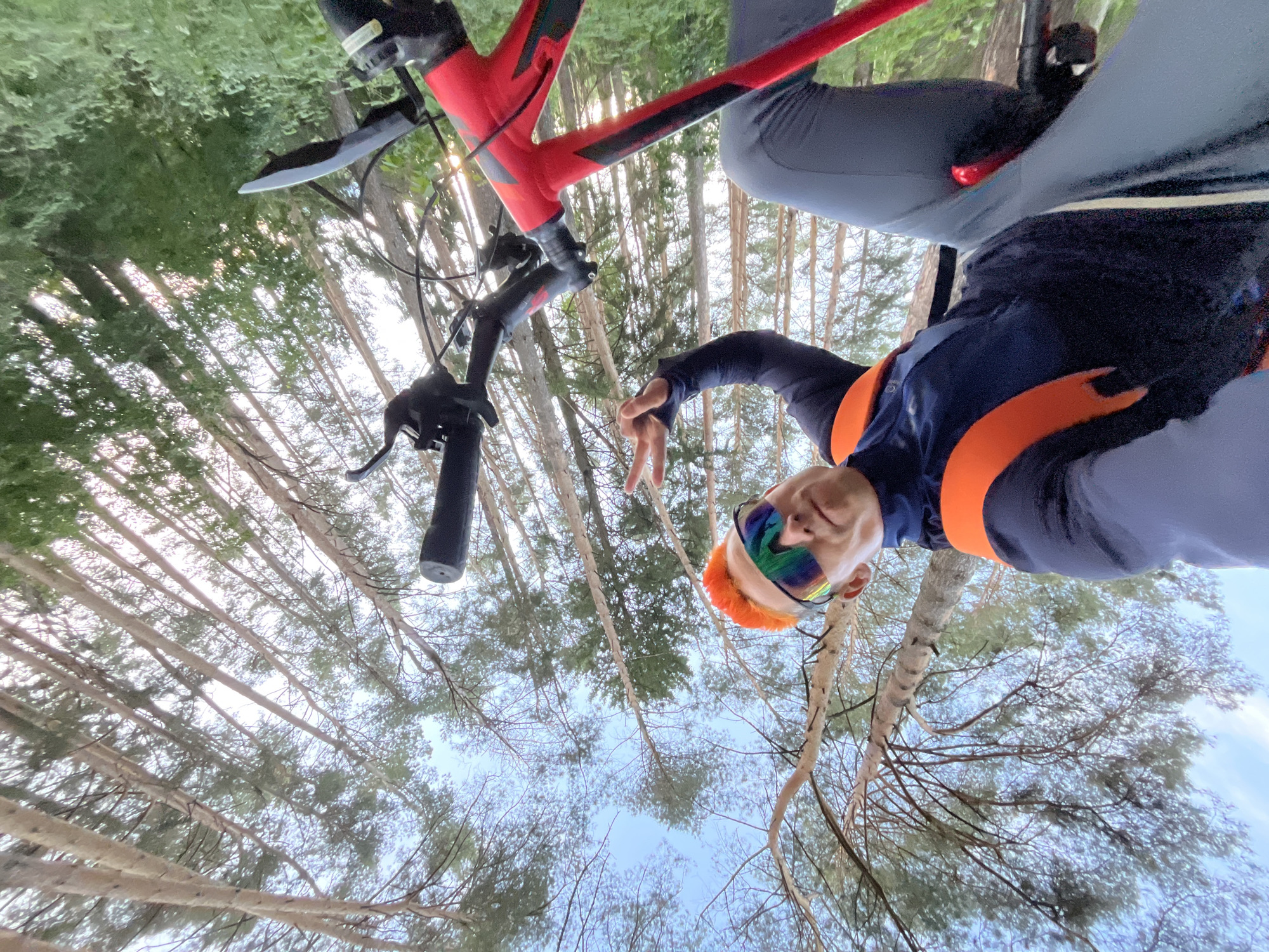The image shows a person with orange hair and colorful reflective sunglasses, wearing a navy blue and orange athletic top and grey pants. They are holding up two fingers in a peace sign. The person is positioned underneath a bicycle with a red frame and black handlebars. The background consists of tall trees with bare branches and a patch of blue sky visible through the canopy. Based on the context provided, the location is assumed to be in the city of Chaikovsky, Russia.