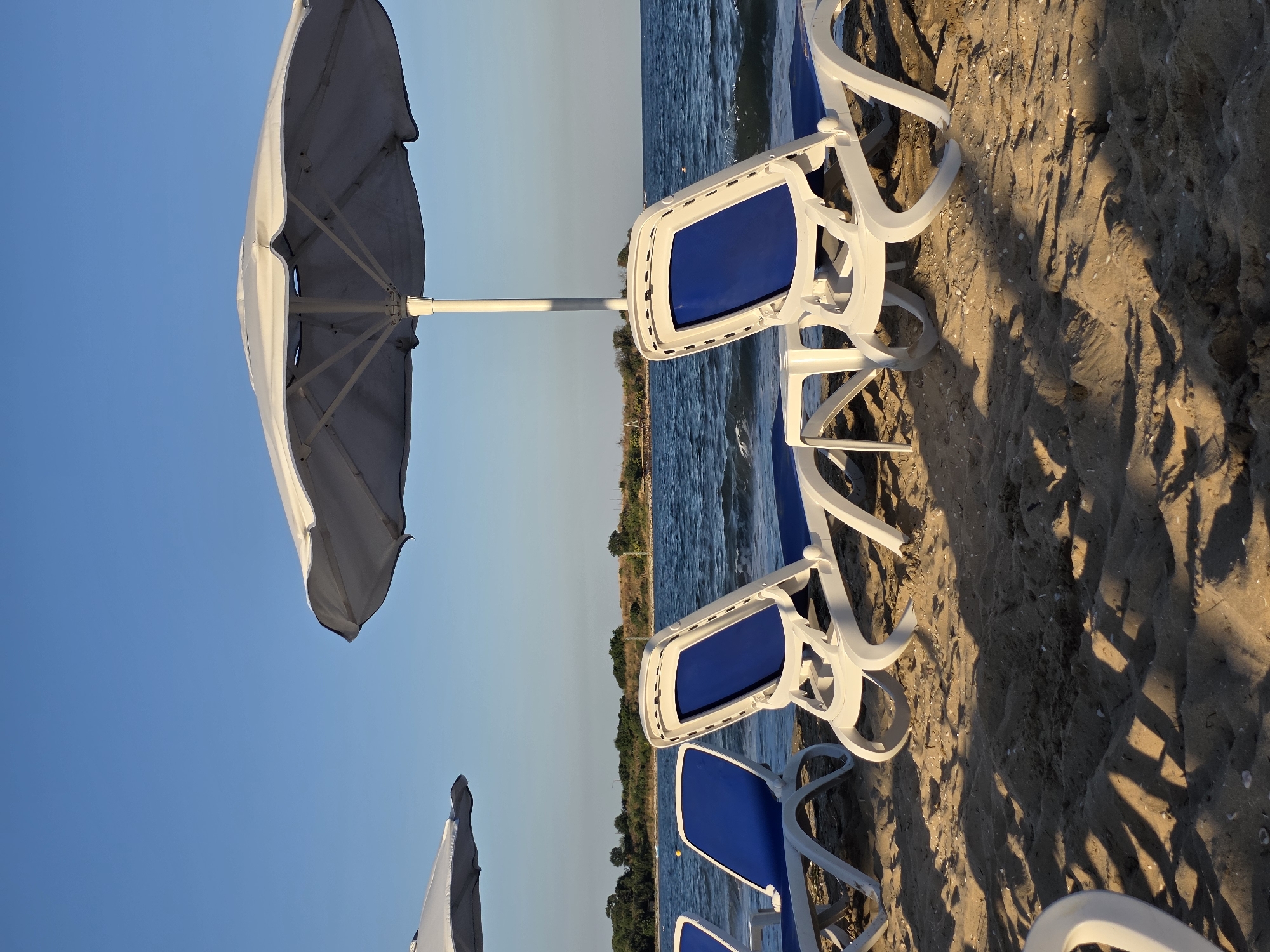This image captures a serene beach scene in Chernomorets, Bulgaria. The foreground is dominated by tan-colored sand, with several white plastic lounge chairs with blue cushions arranged in a row. One large, off-white umbrella stands prominently, casting shadows on the sand, while a portion of another umbrella is visible to the left. Beyond the chairs, the calm, blue sea stretches to the horizon. A strip of green vegetation and trees marks the distant shoreline. The sky is a clear, bright blue. There are no people visible in the immediate foreground or middle ground of the image, although a distant figure can be seen swimming in the sea. The overall impression is one of tranquility and relaxation, indicative of a typical day at the beach in this Bulgarian coastal town.