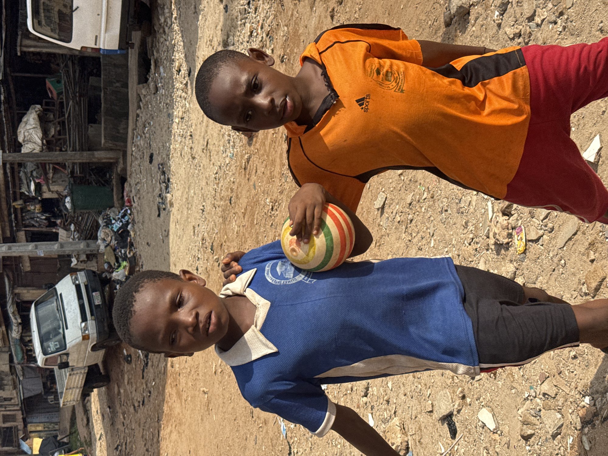 Two boys stand on a sandy, uneven surface in Onitsha, Nigeria. The boy in the foreground wears a blue polo shirt with white trim, black shorts, and holds a multicolored ball. The boy behind him wears an orange Adidas shirt with black stripes and red pants. A white van is visible in the background, surrounded by a cluttered environment with scrap materials and rudimentary structures.