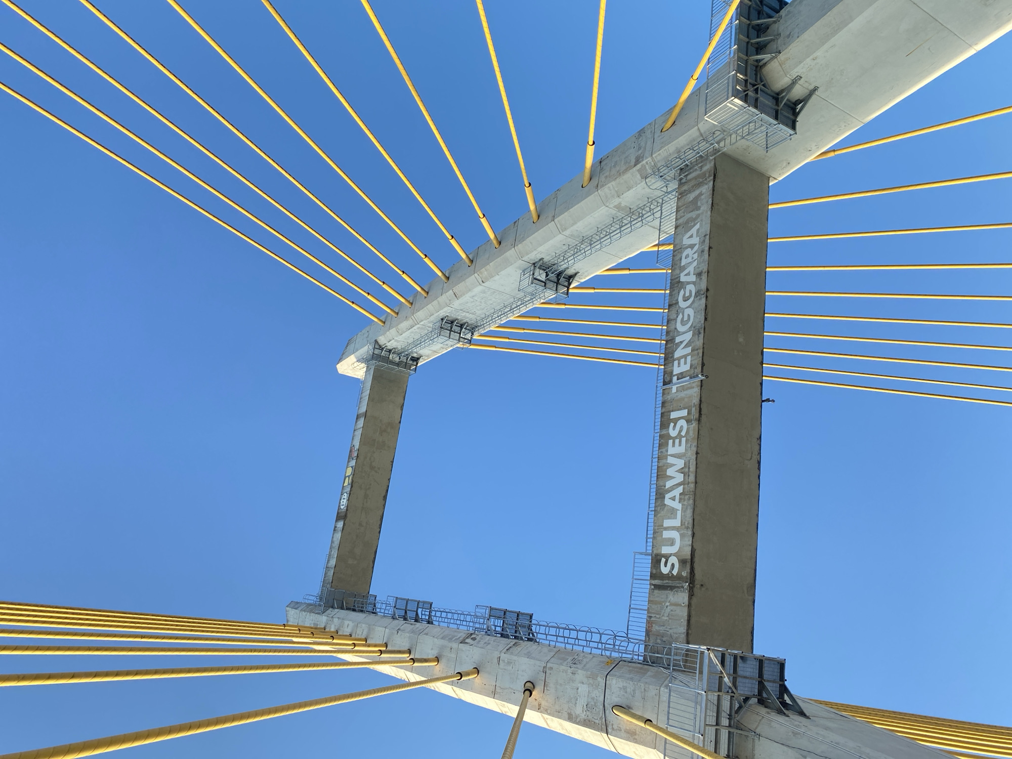 The image captures a low-angle view of a modern cable-stayed bridge under a clear blue sky. The bridge features two massive concrete pylons, one on the left and one on the right, angled towards each other. Each pylon supports a network of bright yellow cables that fan out to hold the bridge deck, which is mostly out of frame.
The pylon on the left is taller and thicker, with a metal ladder attached to its side, leading up to the bridge deck. There are metal platforms and railings at various levels. The pylon on the right is also substantial, with similar yellow cables and access structures.
A horizontal concrete beam connecting the two pylons is visible, and it bears the white lettering "SULAWESI TENGGARA". This suggests the bridge is located in Southeast Sulawesi, Indonesia. There is some graffiti on the concrete structures, including what appears to be "PP" on the upper beam.
The environment is bright and sunny, with no visible people or vehicles. The focus is on the impressive engineering and the bold colors of the bridge against the expansive blue sky. The image is taken from a perspective looking upwards, emphasizing the height and scale of the structure.
