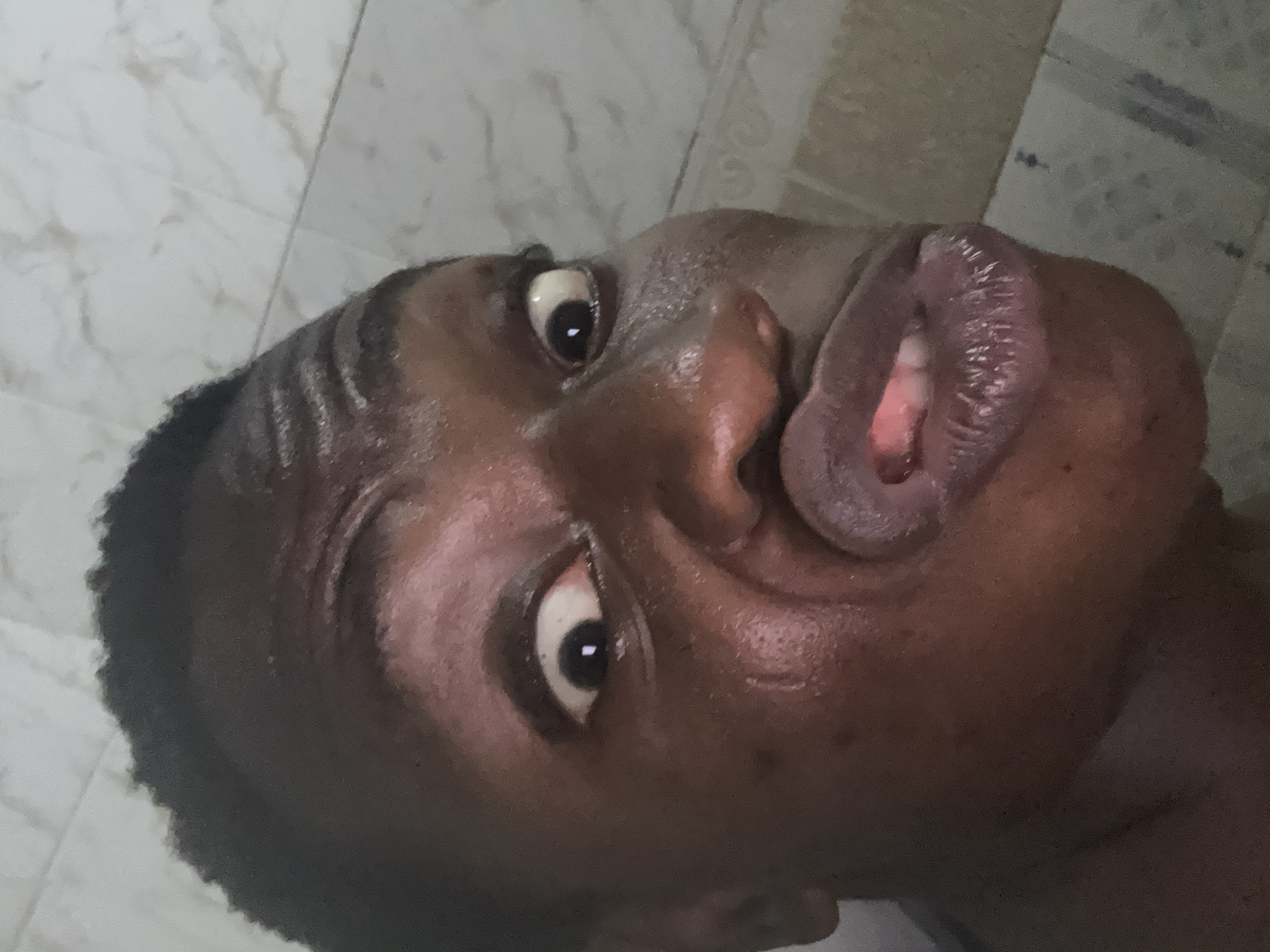 The image shows a close-up, upward-facing perspective of a person's face with exaggerated, pursed lips. The person has dark skin, dark hair with shaved lines, and visible moisture on the skin. The background appears to be a tiled surface, possibly a bathroom wall or floor. No specific city or location details are discernible from the image.