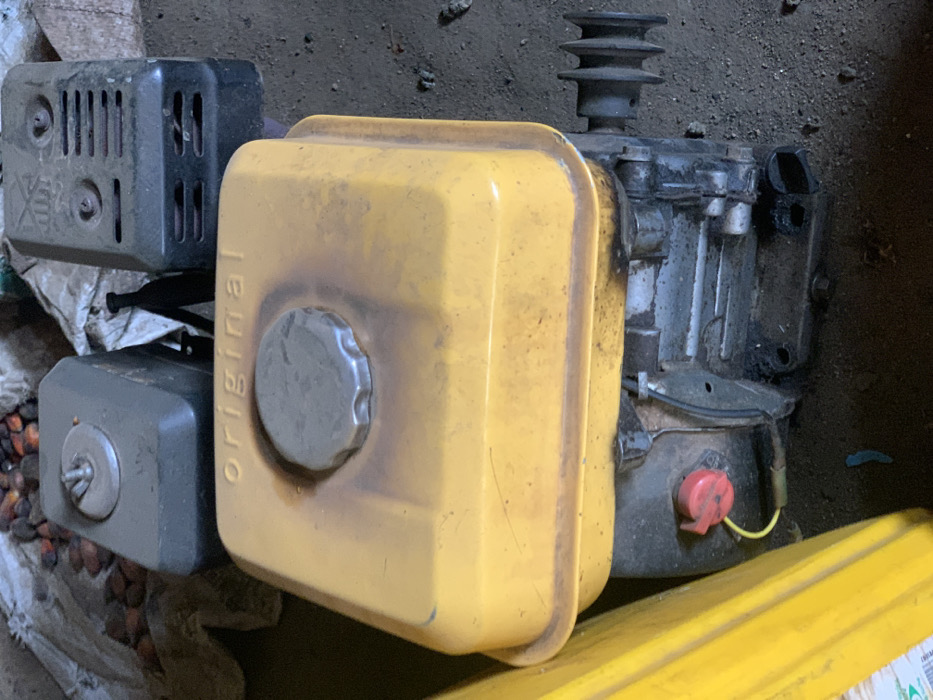 The image shows a small gasoline engine, likely for a generator or other small machinery, lying on the ground. The engine has a yellow fuel tank with the word "ORIGINAL" written on the side. The engine is partially covered by a black plastic cover. The engine is lying on a rough surface that appears to be dirt or gravel. There is a red switch on the engine. The coordinates provided, latitude: 7.12, longitude: 5.362, place the image in the Egbeda area of Lagos, Nigeria. The engine is in a state of disrepair and is covered in dirt and grime. There is no one in the scene. There is a yellow plastic container in the bottom right of the image. The image is likely taken from above, at a slightly tilted angle. There are other objects visible in the image, including a pile of what appears to be palm fruit, but they are not in focus.
