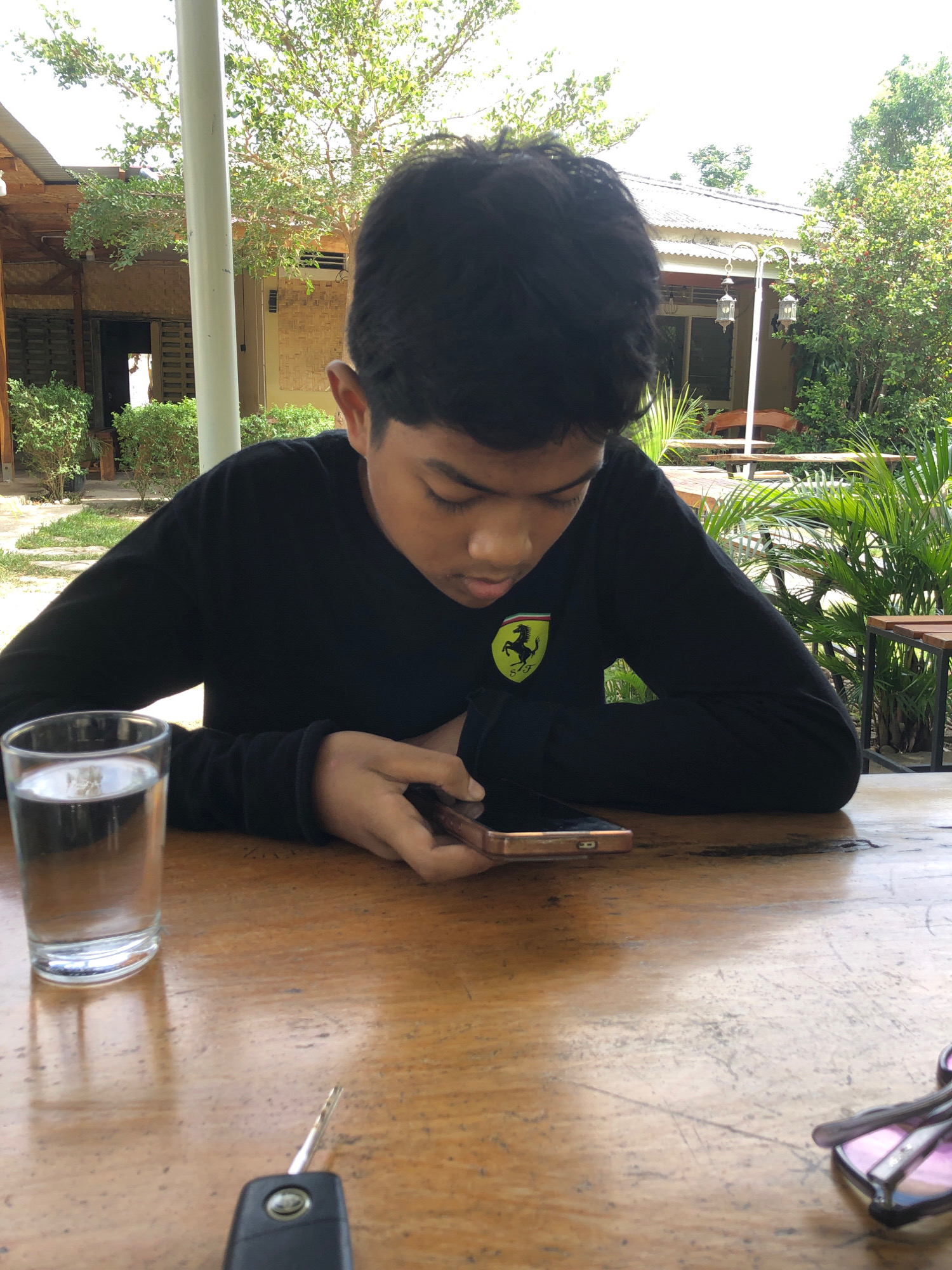 A young male with dark hair is looking down at a gold-colored smartphone in his hands, seated at a wooden table outdoors. He is wearing a black long-sleeved shirt with a yellow and black logo on the left chest. To his left is a clear glass filled with water. In the foreground of the table, a car key fob is visible. In the background, there are green trees, a building with a thatched roof, and outdoor seating areas with tables and chairs. The lighting suggests it is daytime, possibly in Tambonbaroh, Indonesia, given the architectural style and vegetation.