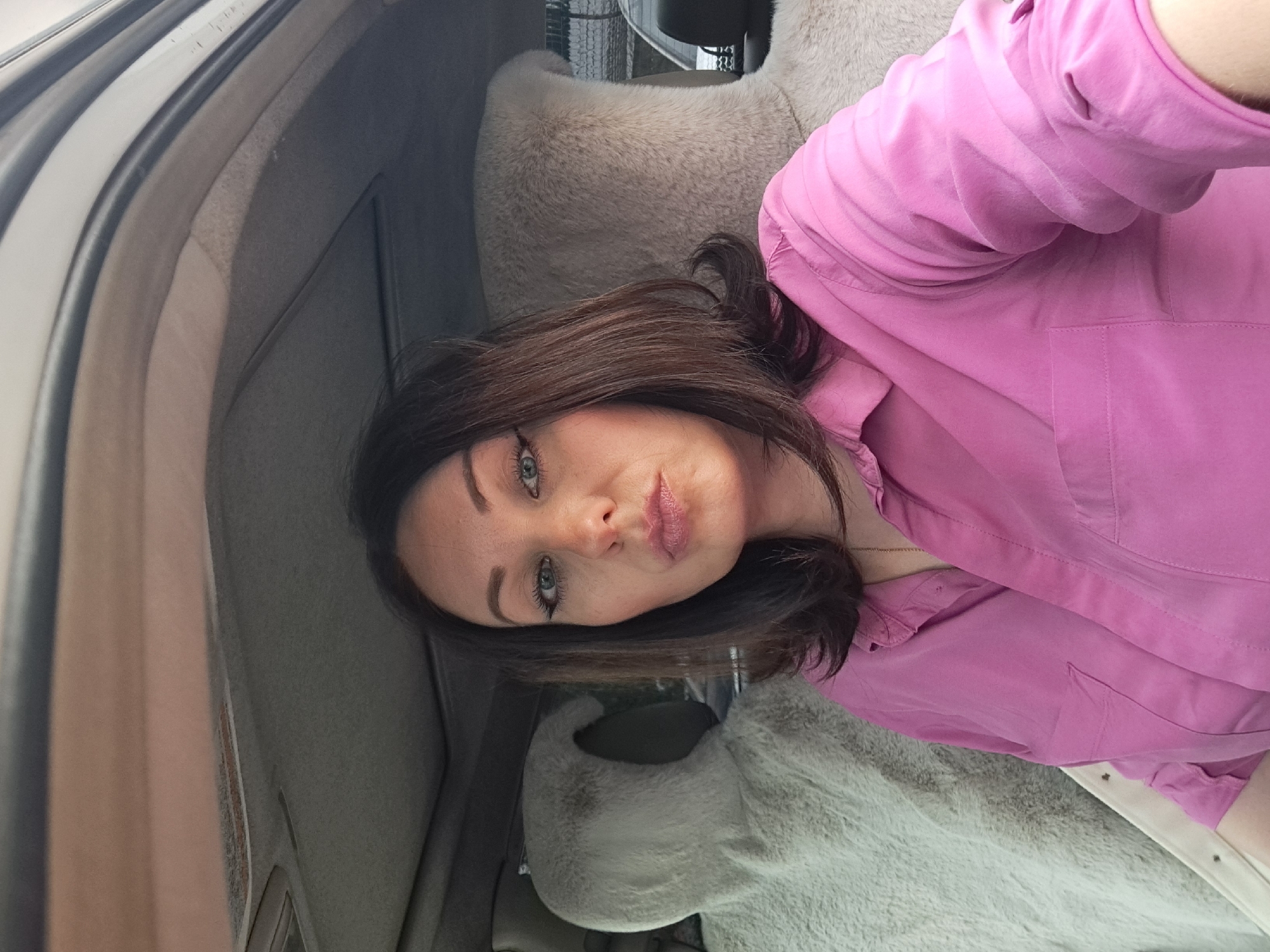 A woman with dark brown hair, blue eyes, and full lips is taking a selfie inside a vehicle. She is wearing a pink collared shirt. The interior of the vehicle is visible, including fuzzy grey seat covers on the headrests of the seats behind her. The sky appears overcast, and there are trees visible in the background, suggesting an outdoor setting. The image was captured in Irkutsk, Russia.