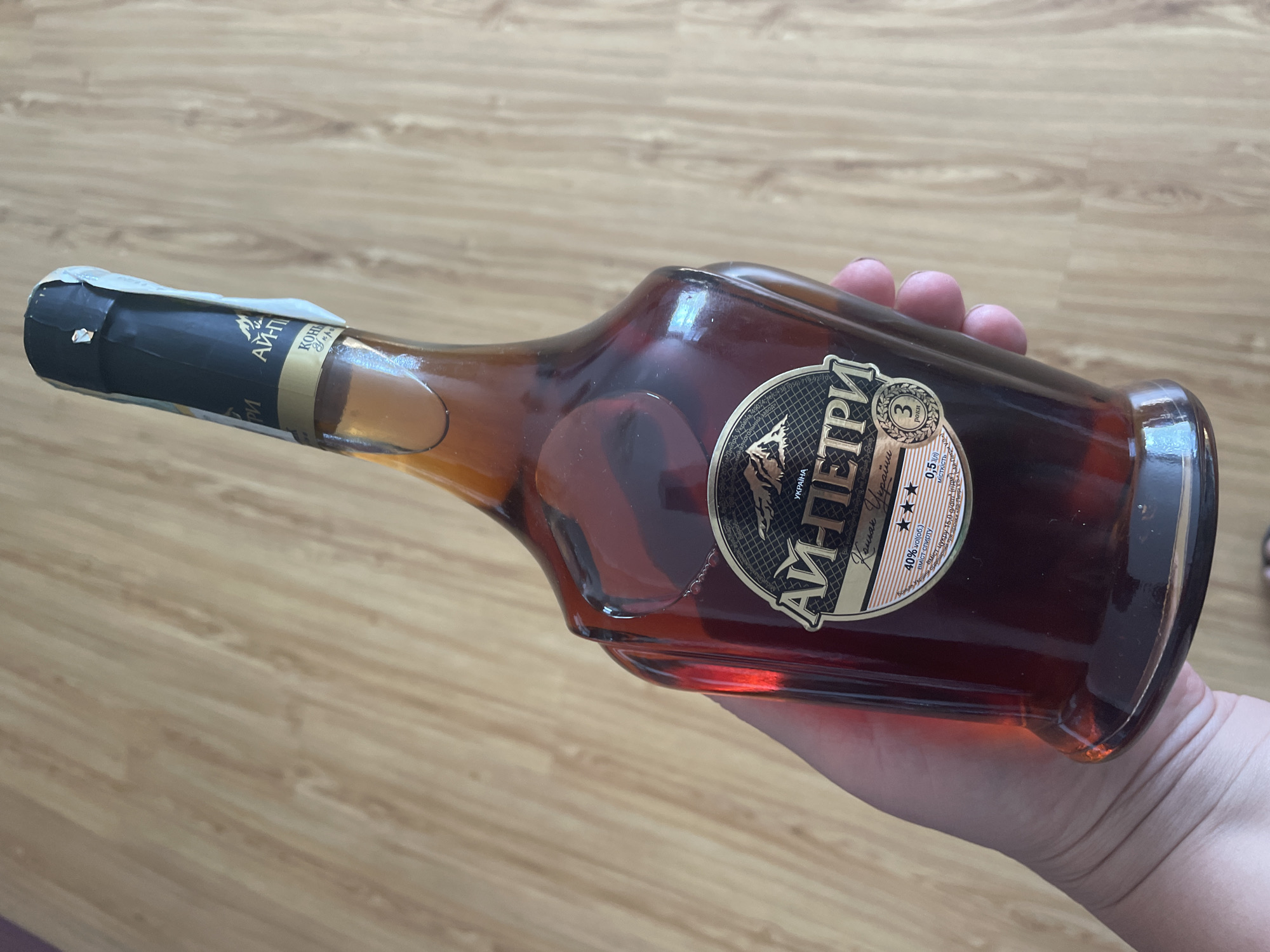 A hand holds a bottle of AI-PETRI cognac on a wooden surface. The bottle is made of brown glass and is tilted to the right. The label on the bottle has a black background with gold lettering and a picture of mountains. The label also indicates that the cognac is 40% alcohol by volume and has been aged for 3 years. The wooden surface appears to be a table or floor. The lighting is natural, coming from above and to the left, casting shadows on the bottle and the hand. The location is not in the city of Shpykolosy, Ukraine, as the bottle's label only indicates "UKRAINA" for Ukraine.