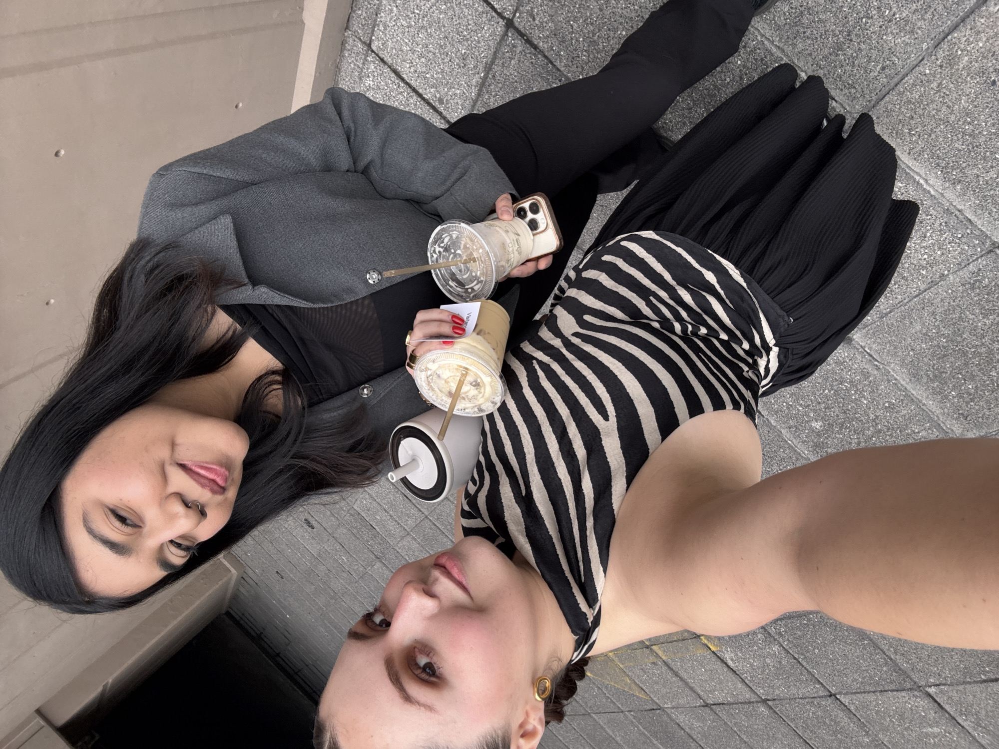 An outdoor selfie, rotated 90 degrees clockwise, features two women posing casually, likely in an urban setting in Mexico City, Mexico. The shot is taken from a slightly elevated perspective, looking down at the subjects and their refreshments.
On the left, a woman with long, dark hair, wearing a grey textured jacket over a black mesh top and black pants, offers a gentle smile to the camera. She has a septum piercing and light makeup. She holds a gold-colored smartphone in her left hand and a clear plastic cup containing an iced beverage with a straw in her right. A small white label with the text "Vienas" faintly visible is attached to her drink.
On the right, a woman, who appears to be taking the selfie, looks directly towards the camera with a calm expression. Her dark hair is mostly pulled back, and she wears a black and white zebra print top, a black pleated midi skirt, and a single gold-colored hoop earring on her visible ear. She holds a white and black reusable insulated cup with a straw.
Both women appear relaxed and the interaction seems friendly. The setting includes grey tiled pavement and a light brown concrete wall in the background, suggesting a sidewalk or outdoor plaza. The natural, soft lighting indicates it is daytime with pleasant weather.