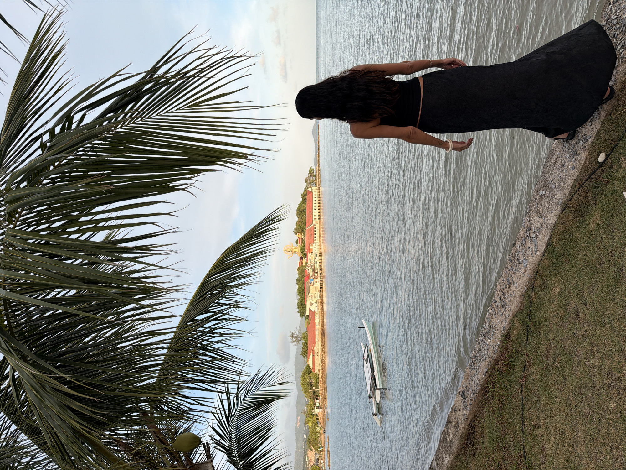 A woman with long, dark hair stands on a grassy bank with a concrete edge, looking out at the water. She is wearing a black tank top and a long, black skirt. In the water, a small, light-colored catamaran is anchored. Across the water, a long, light-colored building with red roofs stretches along the coastline, with hills in the background. A large palm tree extends into the frame from the top left, with a green coconut hanging from it. The sky is a light blue with some wispy clouds. The scene appears to be in the city of Ko Samui District, Thailand.
