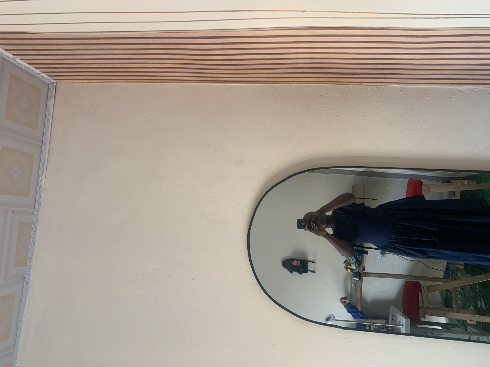 A full-length mirror reflects a person standing in what appears to be a room. The person, wearing a dark blue dress with a tiered skirt, is holding up a phone to take a selfie. Behind the person, in the reflection, is a table with various items on it, including what looks like fabric and a bottle. There are also two red stools visible. The room has plain walls, and one wall has vertical stripes in shades of pink and white. The ceiling has a patterned design. The floor in the reflection appears to be covered in a green, possibly artificial, grass-like material. The overall setting suggests a domestic or workshop environment. There is no indication that this scene is located in Jauro Yinu, Nigeria.