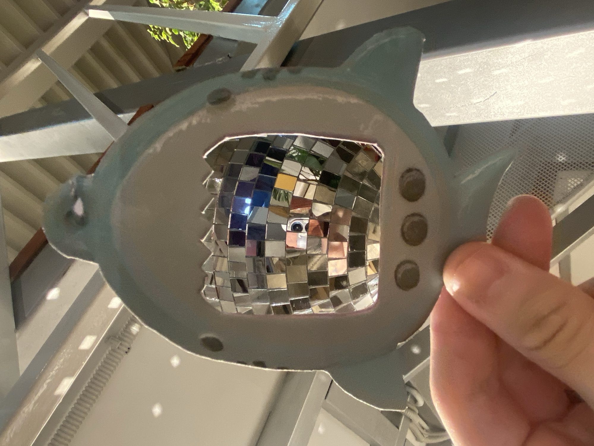 A person's hand is holding a light blue, shark-shaped object. The shark's mouth is open, revealing a mosaic of small mirrored squares that create a distorted reflection of the surroundings and a person's face. The background suggests an outdoor or semi-outdoor setting with architectural elements, possibly a balcony or veranda, with railings and a slatted ceiling. The image is taken from a low angle, looking upwards. The location is not specified in the image or provided context.