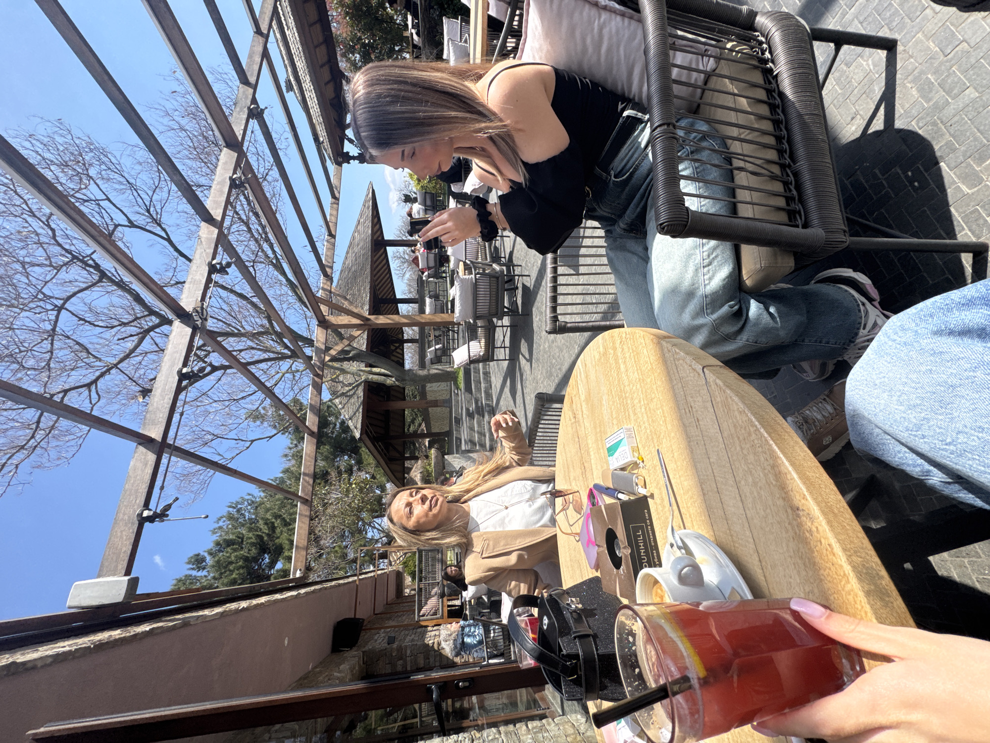A sunny, outdoor scene depicts two women at a table at a cafe, likely in Varna, Bulgaria, given the Dunhill cigarettes on the table indicate a European location, possibly Bulgaria due to the Cyrillic script on the packaging. One woman is seated with her back to the camera, wearing jeans and a black top. She is looking at her mobile phone. Another woman, with blonde hair and a tan jacket, is sitting on the opposite side of the table. A table laden with drinks, a cigarette packet, and personal items is in the foreground. In the background, other tables and chairs suggest an outdoor cafe setting with a trellis structure overhead.