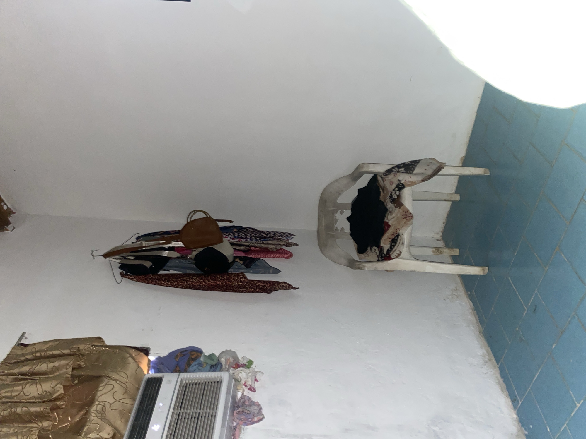 An indoor corner scene shows a white plastic chair in the foreground, overloaded with folded clothing, including a black garment on top and patterned fabrics below. To the left, an air conditioning unit is installed in a wall, with laundry hanging from its frame and on the floor below. On the back wall, a clothes rack holds several garments, including scarves and a brown handbag. The walls are white and textured, and the floor is tiled in blue. The scene appears to be in a domestic setting.