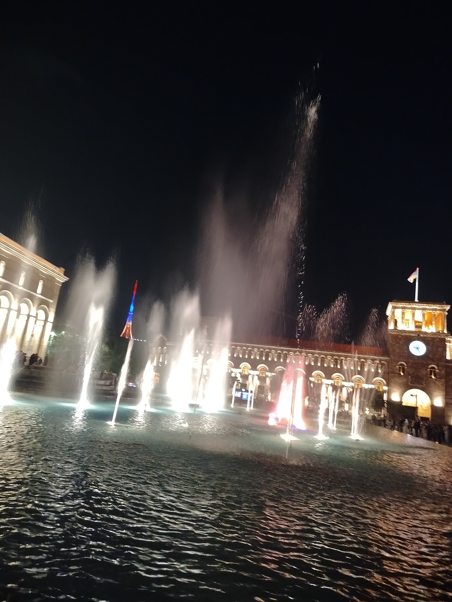 At night in Yerevan, Armenia, a wide pool of water with prominent ripples serves as the base for a fountain display. Multiple jets of water shoot upward, illuminated in white and red, reaching varying heights. In the background, buildings with arched windows and glowing lights line the plaza, with a clock tower visible. A structure featuring the colors of the Armenian flag stands out amidst the water jets. People are seen in the background.