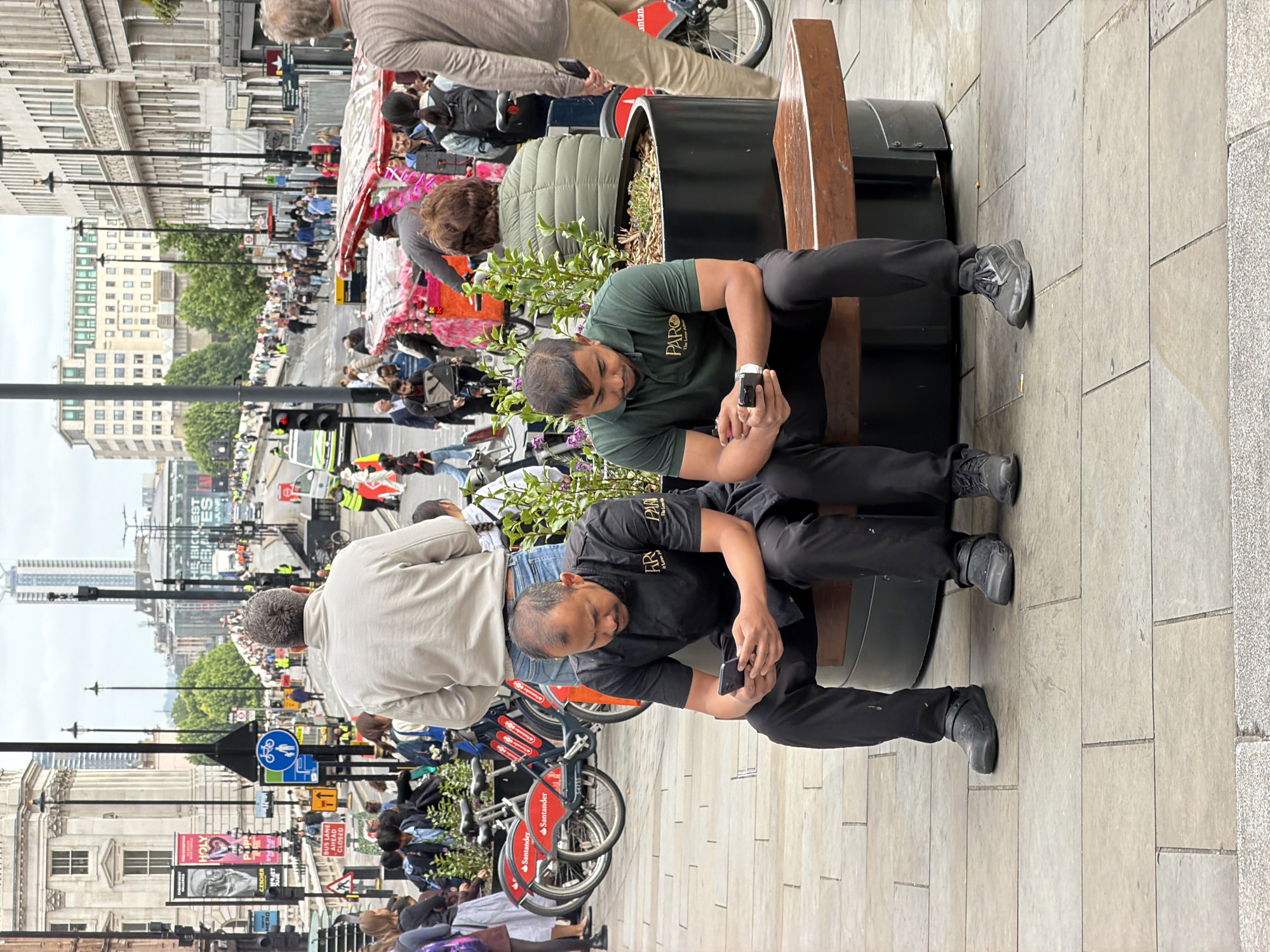 The image depicts a busy urban scene in **Trafalgar Square, London, United Kingdom**, looking towards the east along The Strand or Northumberland Avenue. In the immediate foreground, two men of Asian descent are seated on a communal wooden bench that integrates with a large black planter containing small green plants. Both men are engrossed in their smartphones. The man on the left wears a black chef's jacket with a "PARA" logo, black trousers, and black Crocs-style shoes. The man on the right wears a dark green polo shirt, also with a "PARA" logo, black trousers, and black athletic shoes.
Behind the seated men and to the left, several red Santander Cycles are parked in a row. A man in a light beige jacket and blue jeans stands partially obscured by these bicycles. To the right, another man in a light brown shirt and beige trousers walks away, with a woman in a green puffer jacket seated on a further part of the bench behind the rightmost foreground figure, partially hidden by foliage.
The midground features numerous pedestrians walking along paved footpaths. A colorful pink and orange rickshaw is visible, operated by a man in a black jacket. Street furniture, including lampposts, traffic lights, and various road signs, are scattered throughout; a blue circular sign indicates bicycle and pedestrian routes, while a triangular red sign signifies a give-way junction, and a rectangular sign states "BUS LANE CLOSED."
In the background, a large neoclassical white building with columns, identifiable as **Canada House**, stands prominently on the left, adorned with banners displaying "HOLY" and "ESCHER." Beyond this, a wide street ascends, bustling with a dense crowd of pedestrians and some vehicles, including a white van. Several individuals in high-visibility yellow vests are present among the crowd, potentially marshaling an event. Modern office buildings line the right side of the street, and a tall, slender skyscraper is visible in the distant background under an overcast sky. The ground consists of light-colored rectangular paving slabs.