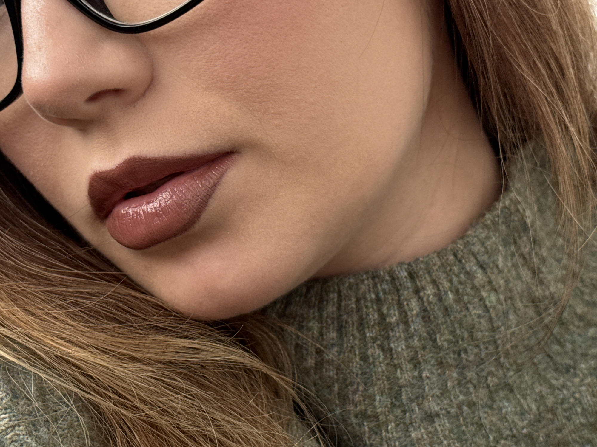 Close-up image of a person's face, focusing on the nose, mouth, and chin. The person is wearing black glasses. The lips are covered in dark brown lipstick with a glossy finish. The person has light brown hair and is wearing a green knit sweater. The location is Unknown, Unknown.