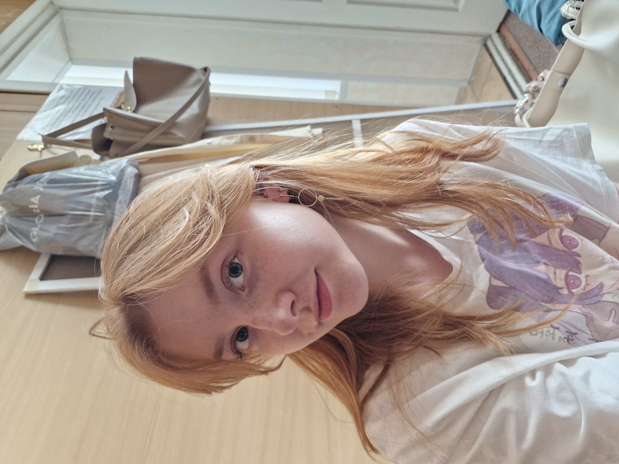 A young woman with reddish-blonde hair and freckles is looking directly at the camera with a slight smile. She has blue eyes. She is wearing a white t-shirt with a cartoon print. To her left, there's a beige purse. Next to the purse is a plastic-wrapped object, and a white frame. The background includes a white shelf or ledge. The location cannot be determined to be Yekaterinburg, Russia, based solely on the visual information provided.