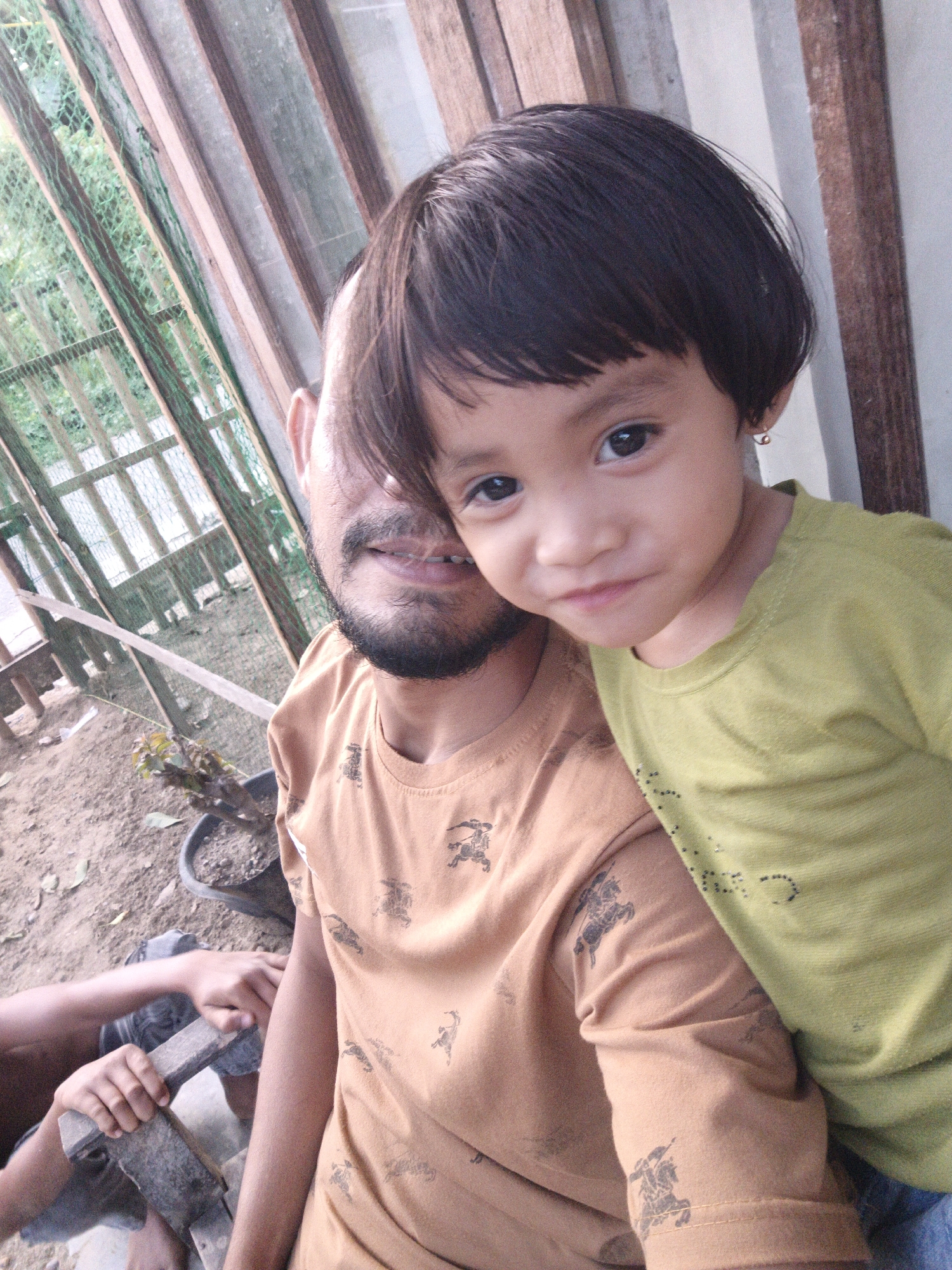In Lhokseumawe, Indonesia, a father and child are captured in a close-up selfie. The father, with a beard and a fringe of hair partially covering his face, wears an orange t-shirt with a repeating pattern of horses and riders. To his right, a young child, likely his daughter, leans into the frame, her face turned towards the camera. She has dark hair and wears a light green t-shirt. The background suggests an outdoor setting, possibly a backyard or rural area, with wooden structures, fencing, and some vegetation visible. The ground appears to be dirt, with a potted plant and loose leaves scattered around. The overall scene conveys a sense of intimacy and connection between the father and child.