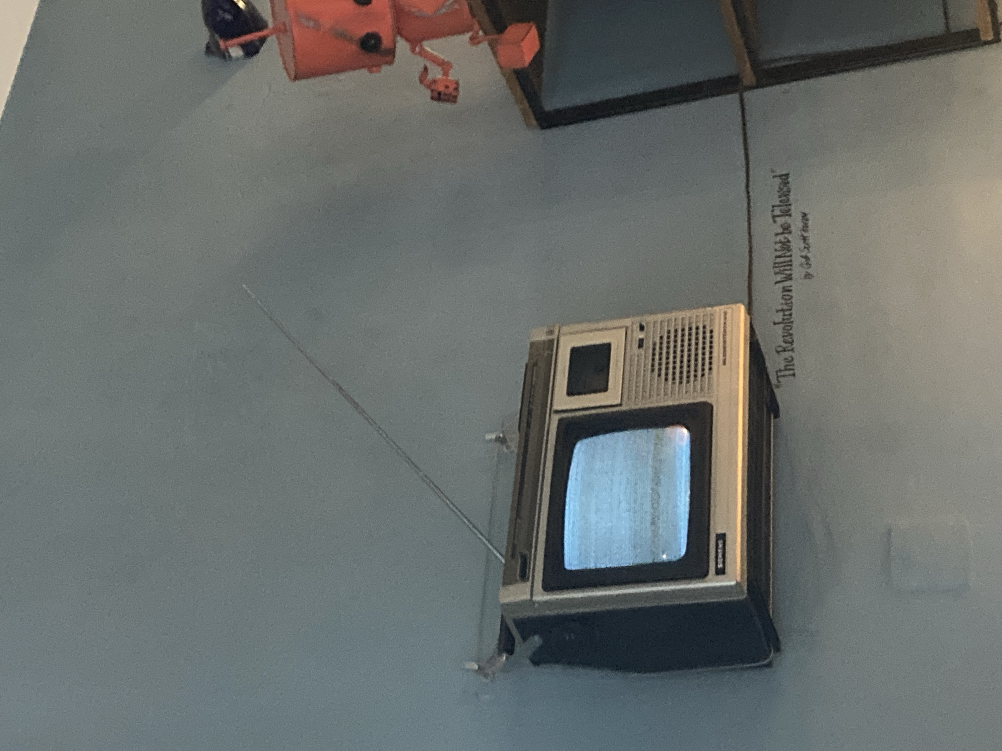 An old-fashioned television is mounted on a blue-grey wall with a retractable antenna extended. To the right of the TV is a framed poster that appears to read, "The Revolution Will Not Be Televised". Above the TV, at the top of the frame, is a piece of what looks like a red and orange robotic or mechanical sculpture. The scene appears to be in an indoor location, possibly a gallery or an art installation, in Sofia, Bulgaria.
