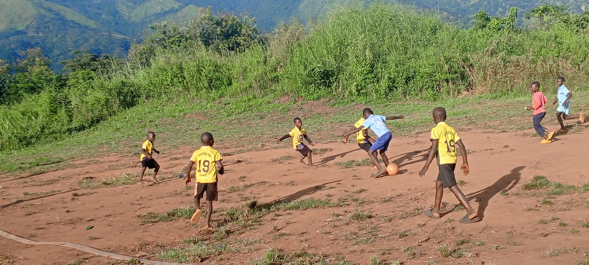 A group of children are playing soccer on a dirt field in Ngalula, Democratic Republic of the Congo. The boys wear varying clothing; some wear yellow soccer jerseys, one wears a blue jersey, and another a red one. A soccer ball is in play. In the background are rolling hills covered with dense vegetation. A gray pipe snakes along the lower-left side of the scene.