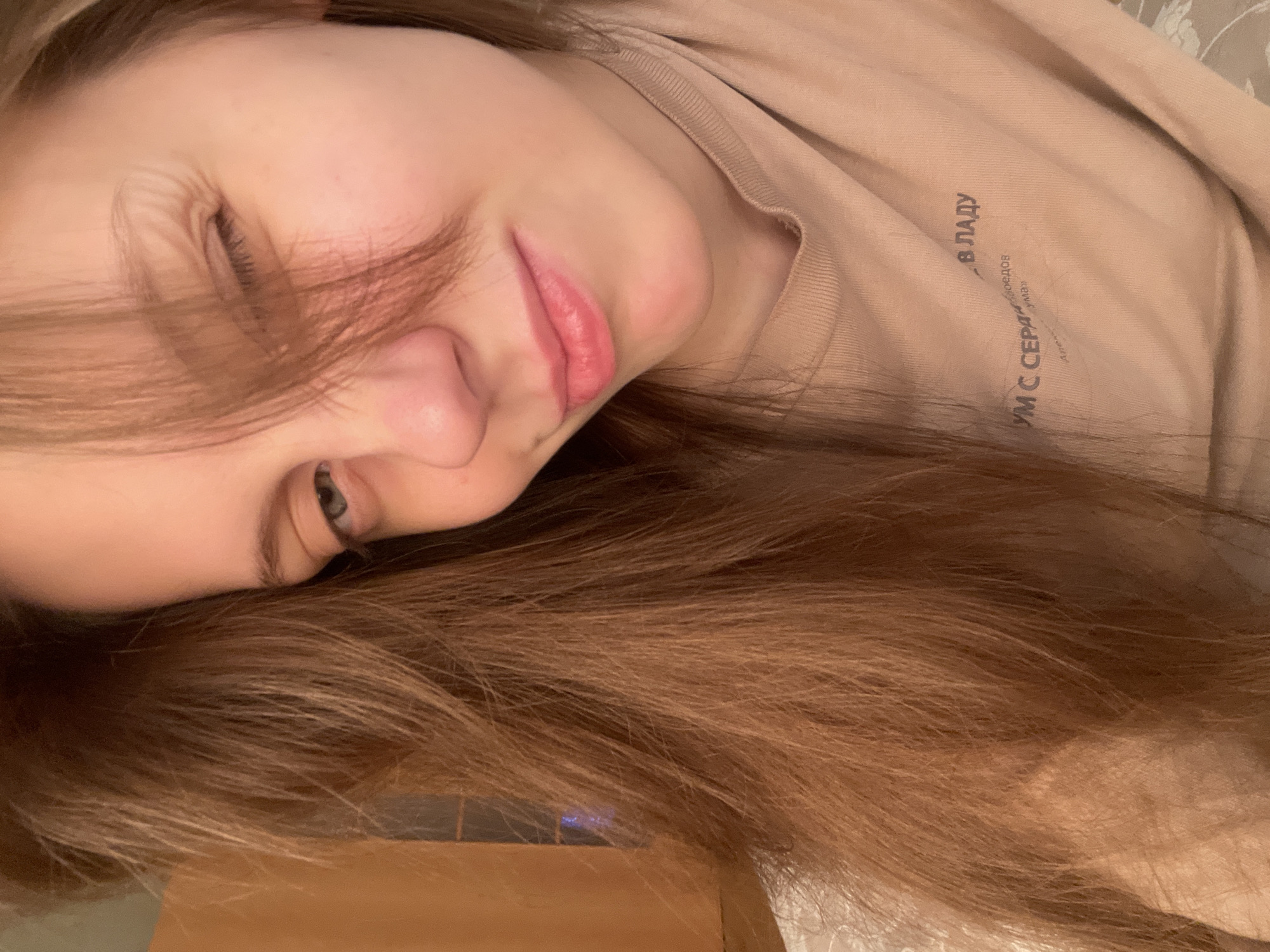 The image is a close-up, selfie-style photograph of a young woman with long, light brown hair. She is winking with her right eye, while her left eye is partially visible. Her hair is falling across her face, obscuring part of her forehead and left eye. She is wearing a light brown t-shirt with text printed on it. The text is in Russian and appears to be a quote or phrase. The background is blurred and indistinct, but suggests an indoor environment with warm lighting. There are no specific landmarks or details in the image that would indicate a location in Ufa, Russia, or any other city.