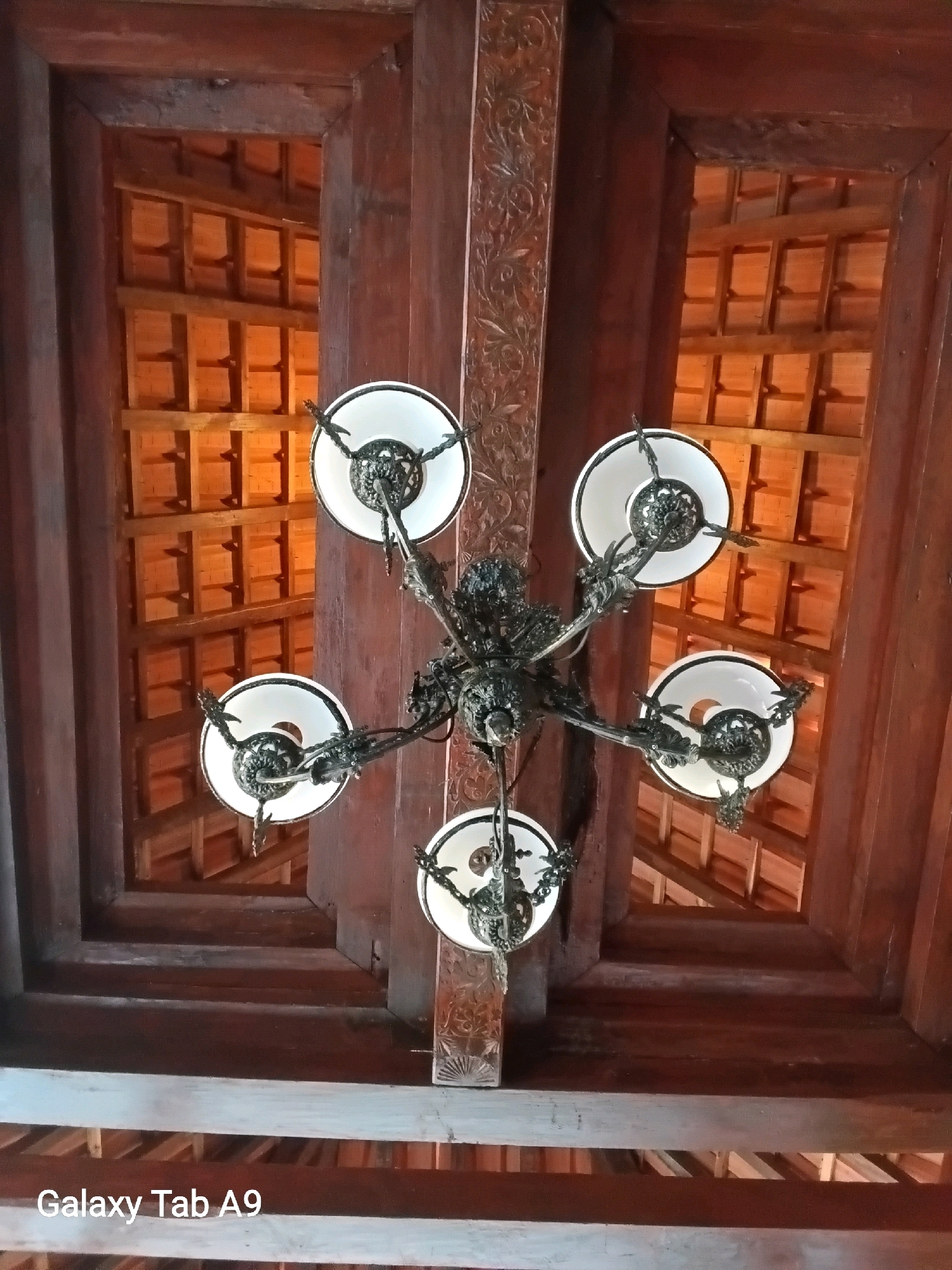 This is a low-angle, close-up, full-shot photograph of an ornate, dark metal chandelier with five white bell-shaped lamps. The chandelier is suspended from a wooden ceiling with visible beams and decorative carved panels. Through openings in the wooden ceiling structure, a sloped roof made of terracotta tiles is visible. The image was taken indoors, likely in a building with traditional architecture. There are no people in the image. The location is identified as Trenggalek, Indonesia.