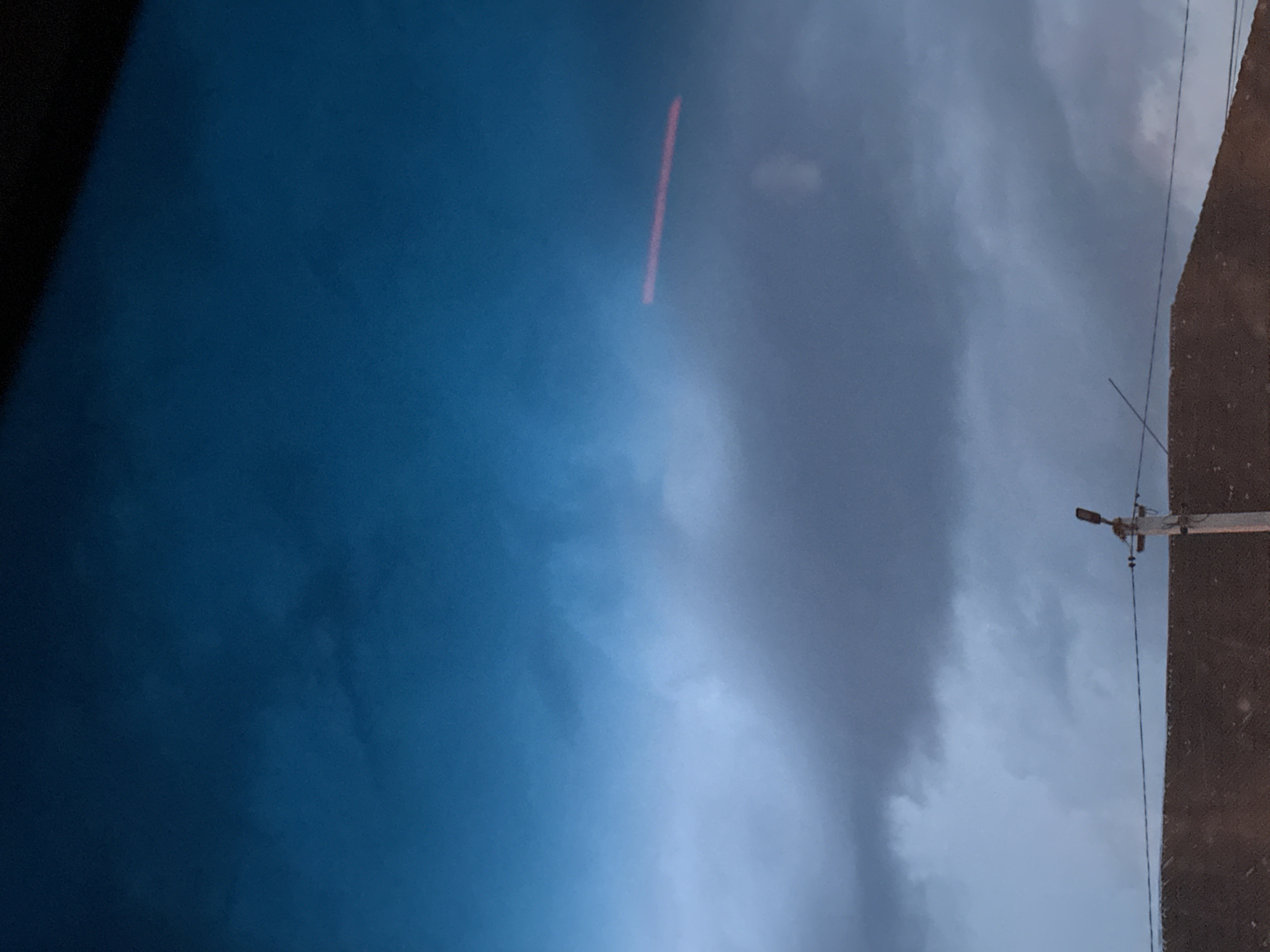 The image depicts a cloudy sky with a gradient from dark blue at the top to lighter blue and grey towards the bottom. A faint red horizontal line is visible in the upper half of the sky. In the lower portion of the image, the top of a brown, textured surface, possibly a rooftop, is visible, with a utility pole and wires extending across. The scene is overcast and appears to be taken at dusk or during a storm. The location is Shatava, Ukraine.