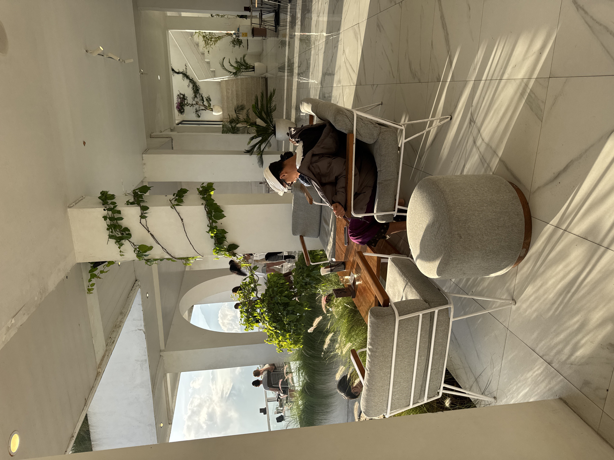The image captures an interior scene in Kedisan, Indonesia. An individual wearing a head covering and a dark coat is seated in a light-gray chair with white metal legs. The chair is positioned at a wooden table. Next to the chair is a round, gray ottoman. Other people are seated at the table, surrounded by decorative plants and foliage. The background features an open-air architecture with white walls, archways, and views of the sky. The floor is tiled with a white marble pattern, and sunlight is cast upon it. The setting appears to be a modern, open-air cafe or lounge.