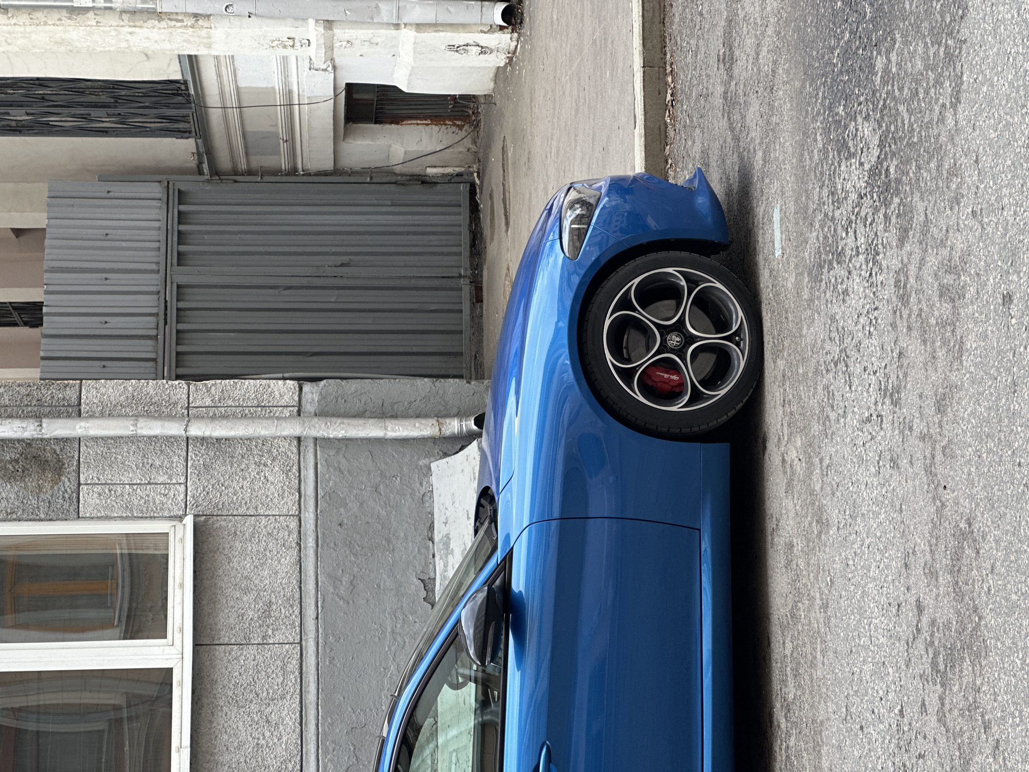 The image captures a portion of a blue car, specifically an Alfa Romeo, parked on a street. The vehicle's front wheel and part of the front section are visible. The wheel has a distinctive five-circle design, with red brake calipers behind it. The surrounding environment includes a gray concrete wall with a window and a metal gate. The street is paved with gray asphalt. The location is Moscow, Russia.