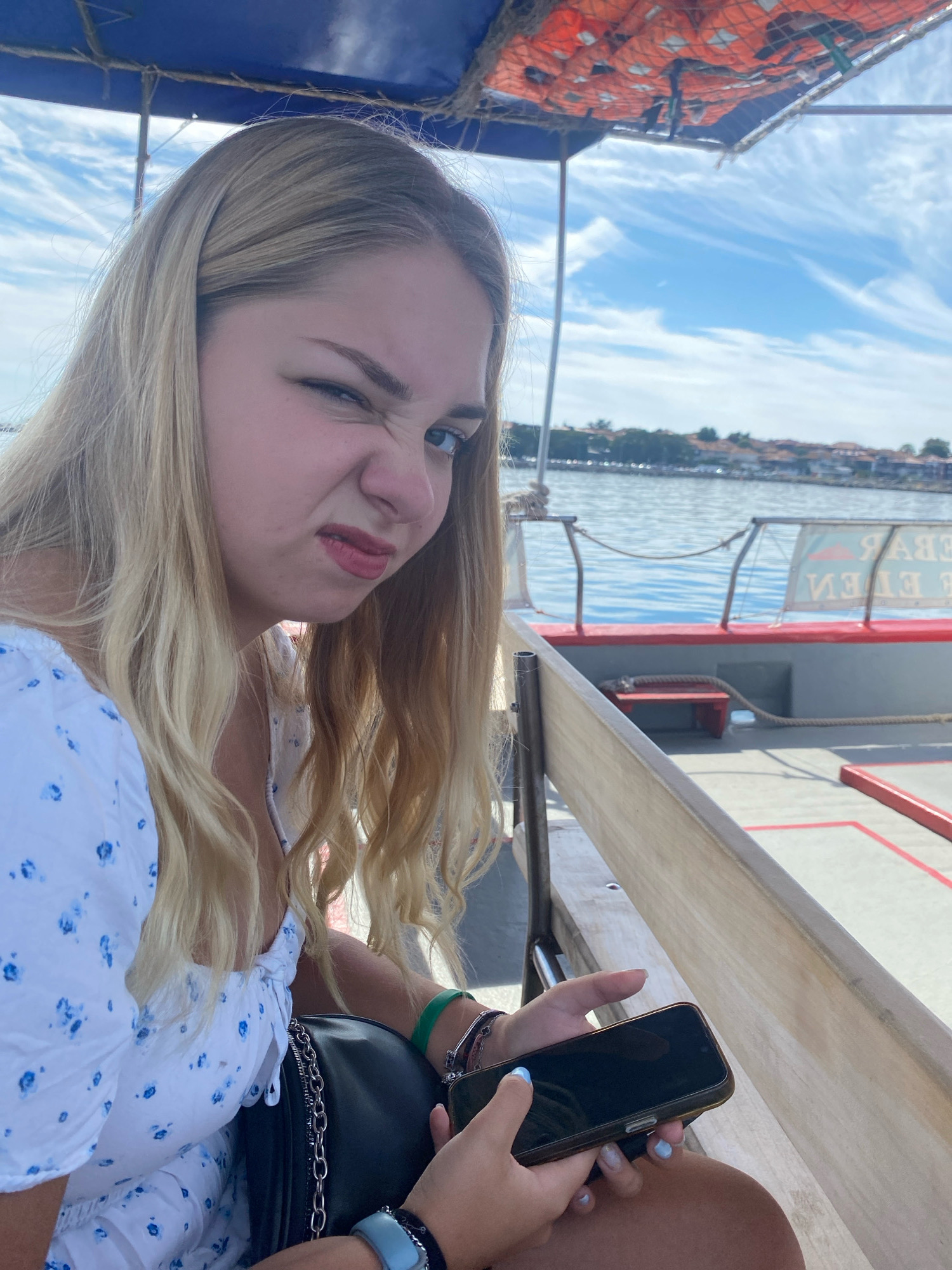A young blonde woman is sitting on a boat, making a disgusted face at the camera. She is wearing a white dress with blue flowers and holding a smartphone. In the background, there is the sea and the city of Nesebar, Bulgaria, with its historic buildings visible along the coastline. The boat has orange life jackets stored under a blue canopy.