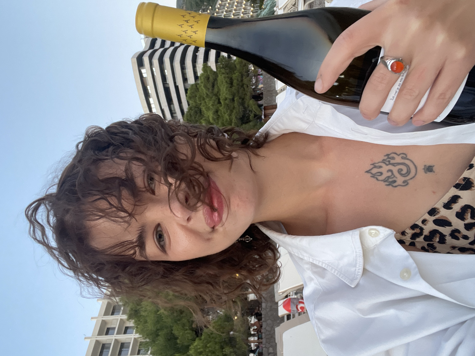 This is a selfie taken outdoors in Rafailovići, Montenegro. A young woman with curly brown hair is holding a bottle of wine. She is wearing a white button-up shirt over a leopard-print top and has a tattoo on her chest. Behind her, there are trees, beach chairs, and modern buildings, suggesting a coastal resort area. The sky is clear and blue.
